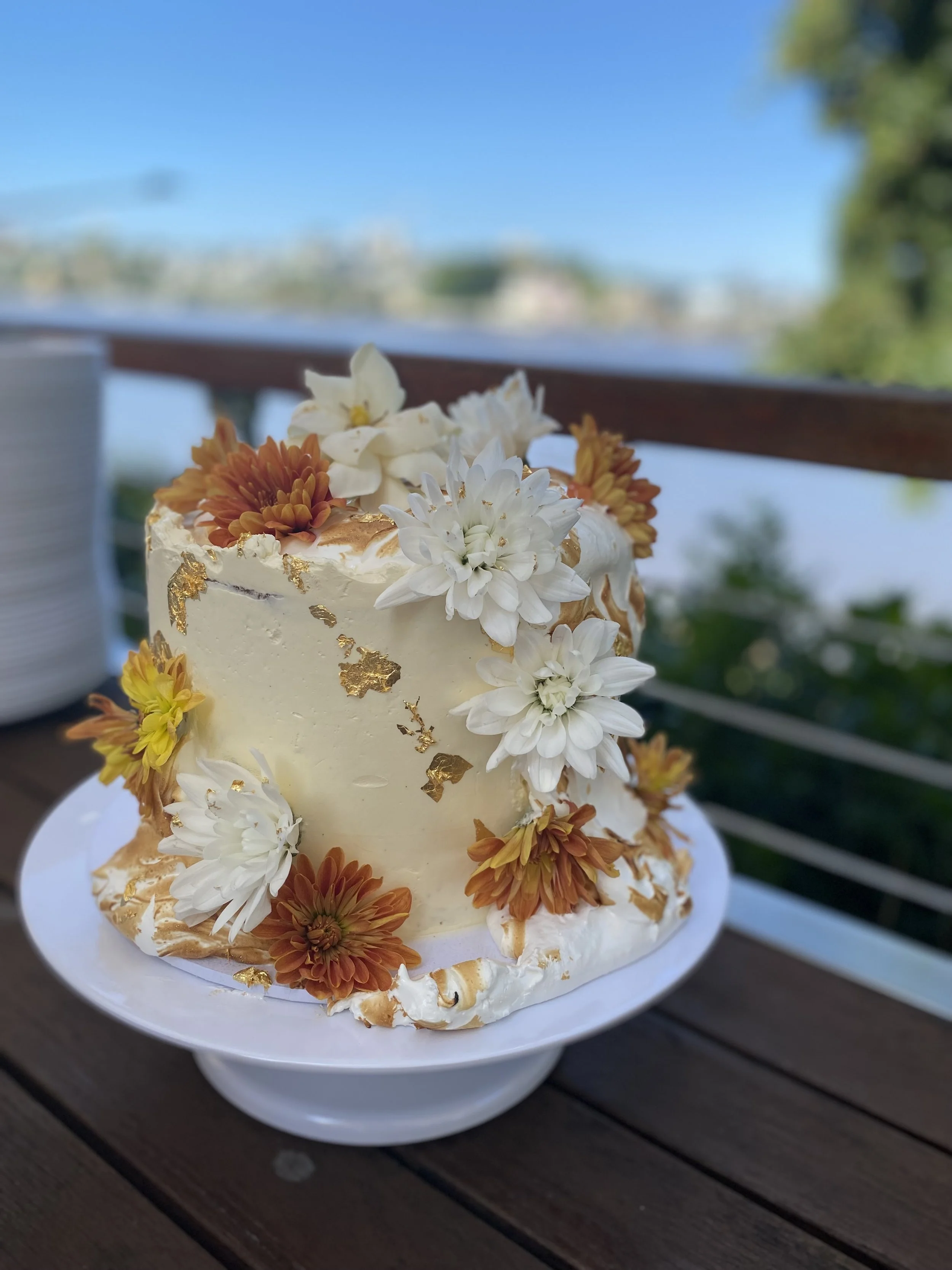 A white frosted cake decorated with white and orange flowers, gold leaf accents, and placed on a white cake stand outdoors on a wooden table with a water body and trees in the background.