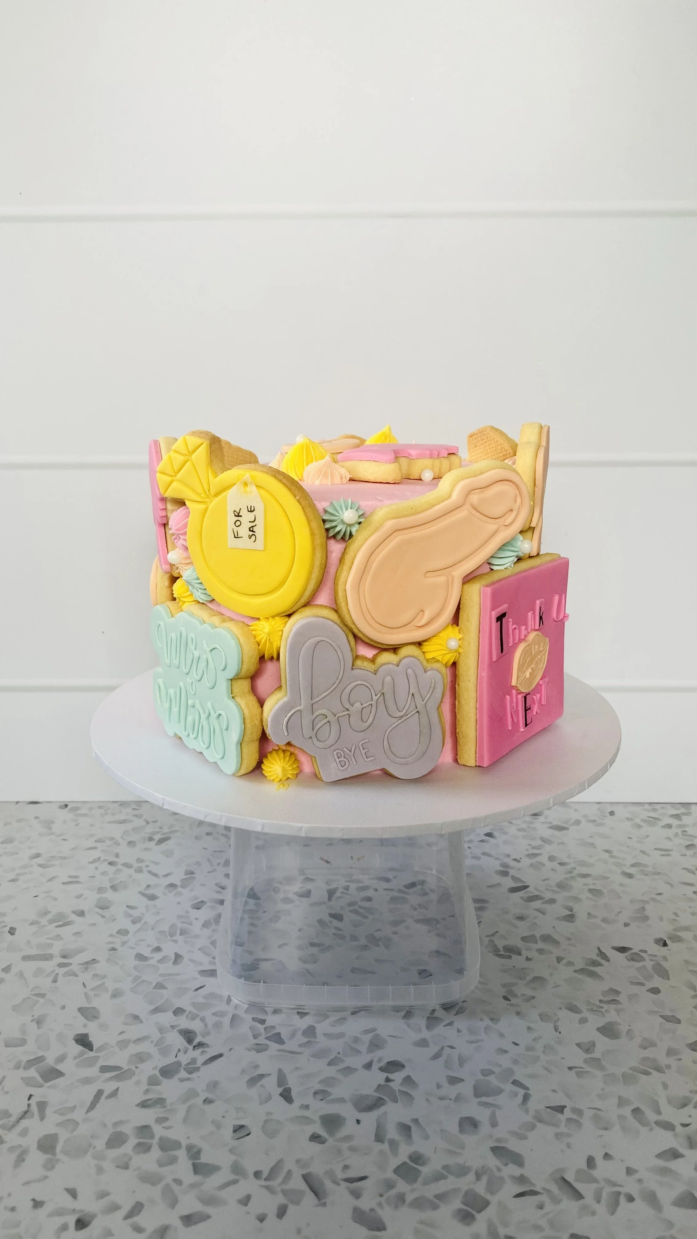 Decorated birthday cake with pastel-colored cookies and icing, featuring phrases like "Bye" and "Thank You," placed on a white cake stand on a terrazzo countertop.