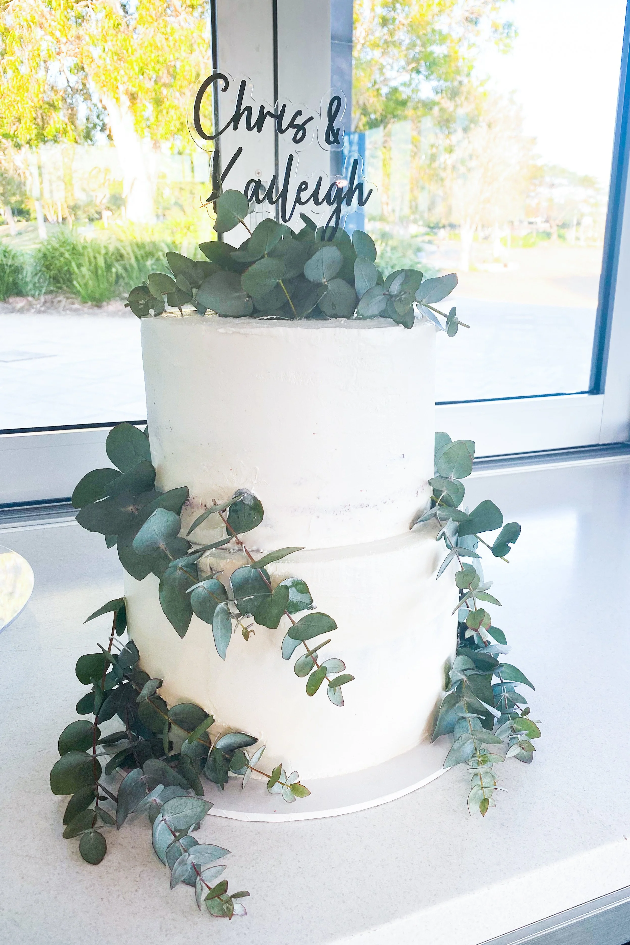 Two-tiered white wedding cake decorated with eucalyptus leaves and a cake topper with the names 'Chris & Kathleen' in black cursive letters, placed on a white surface near a window with trees visible outside.