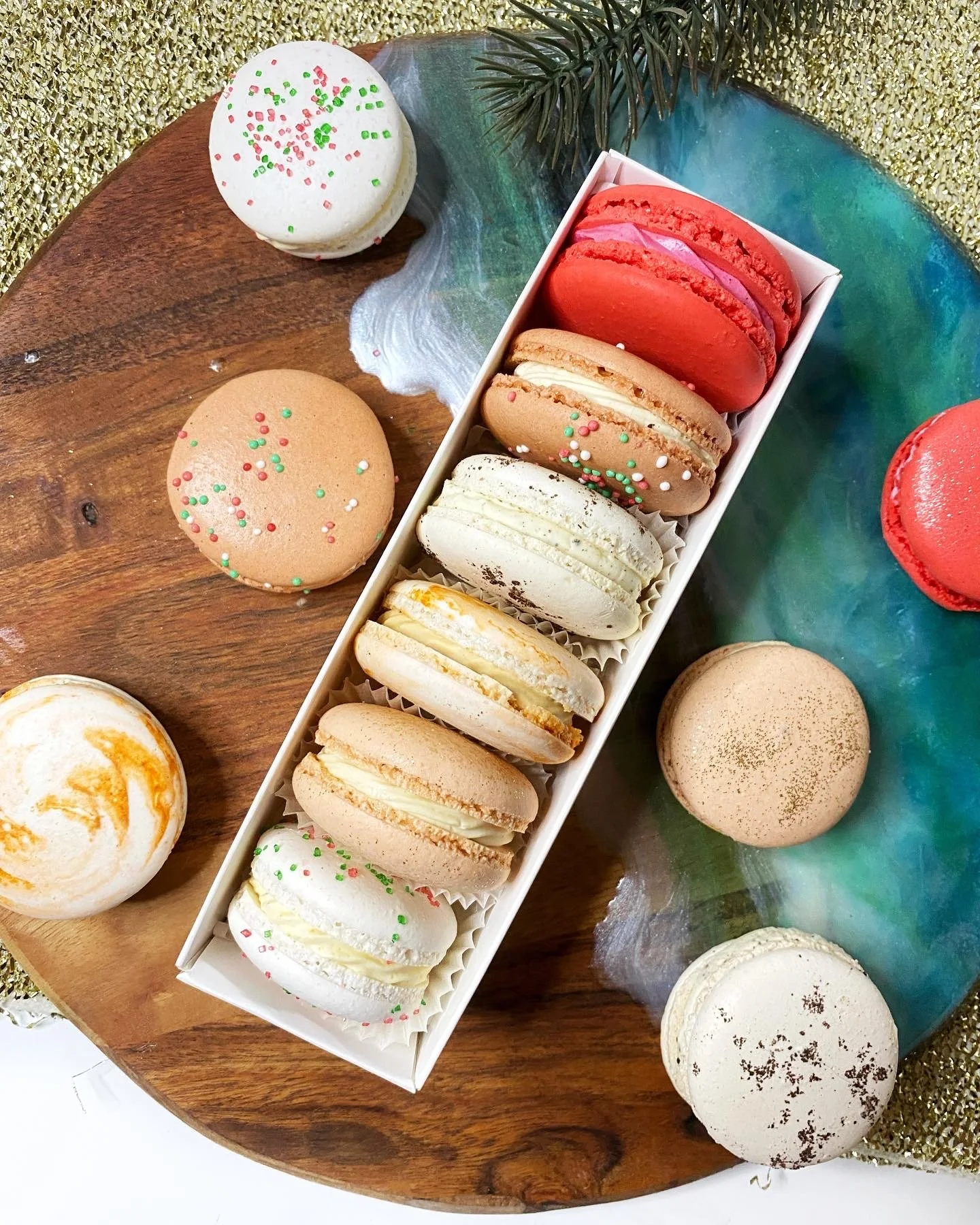 Assorted macarons in a white box on a wooden and colorful surface with sprinkles and a small pine branch.