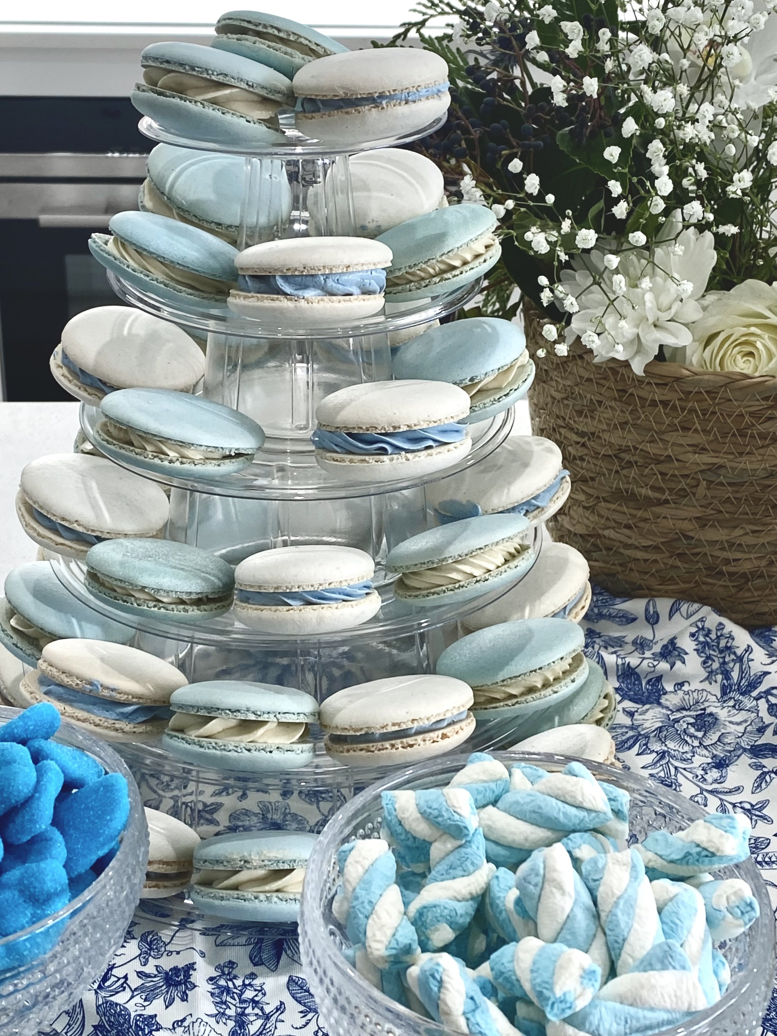 A tiered stand filled with blue and white macarons, decorated with cream filling. To the right, a basket with white flowers and greenery. In the foreground, bowls of blue candy and swirled striped candies on a floral cloth.