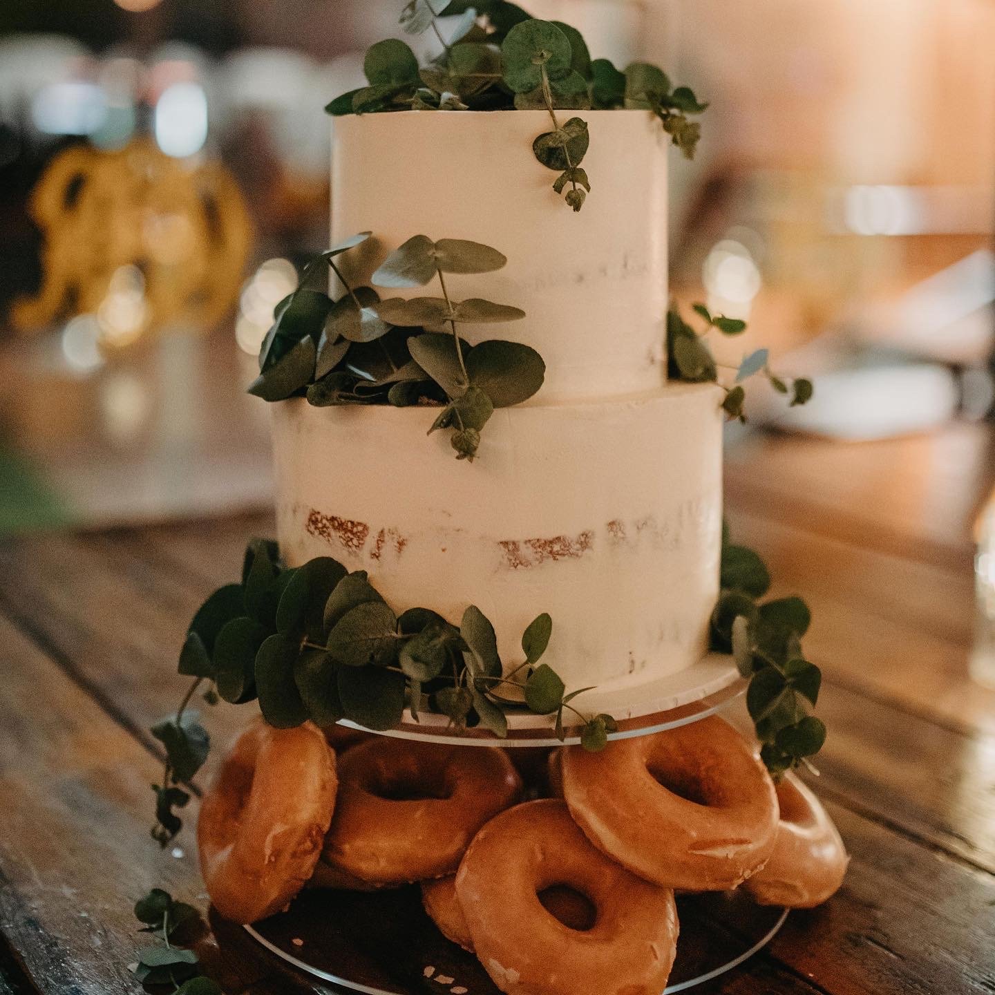 A two-layer white cake decorated with green leaves, placed on a wooden surface surrounded by glazed donuts.