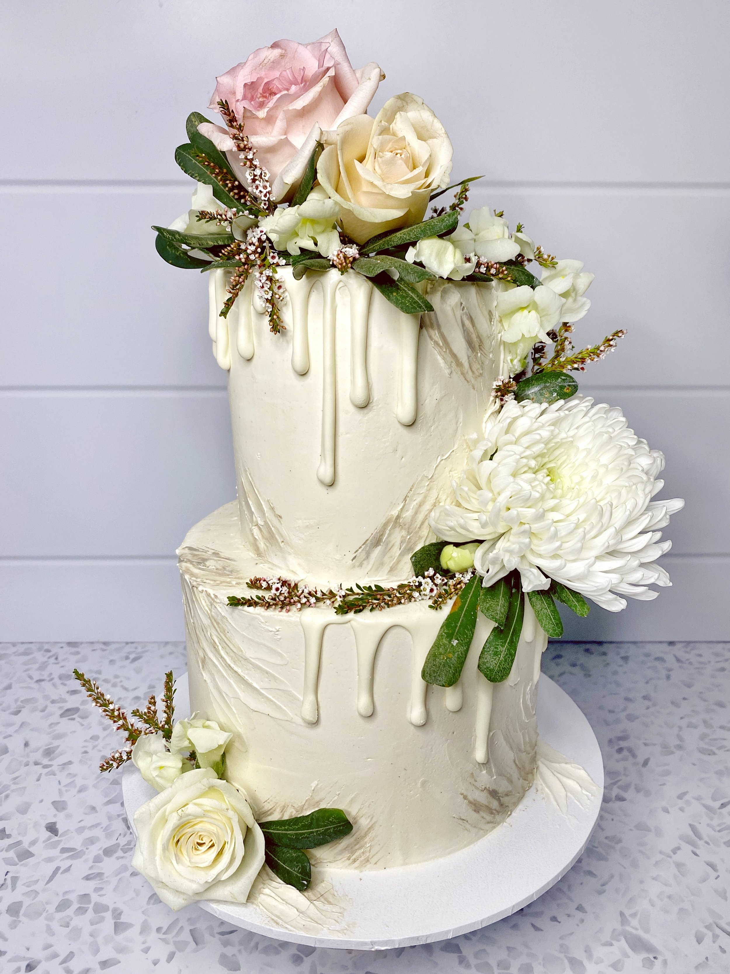 Two-tier white wedding cake with floral decorations and white drip icing, topped with pink and cream roses, white chrysanthemum, and greenery, on a white cake board.