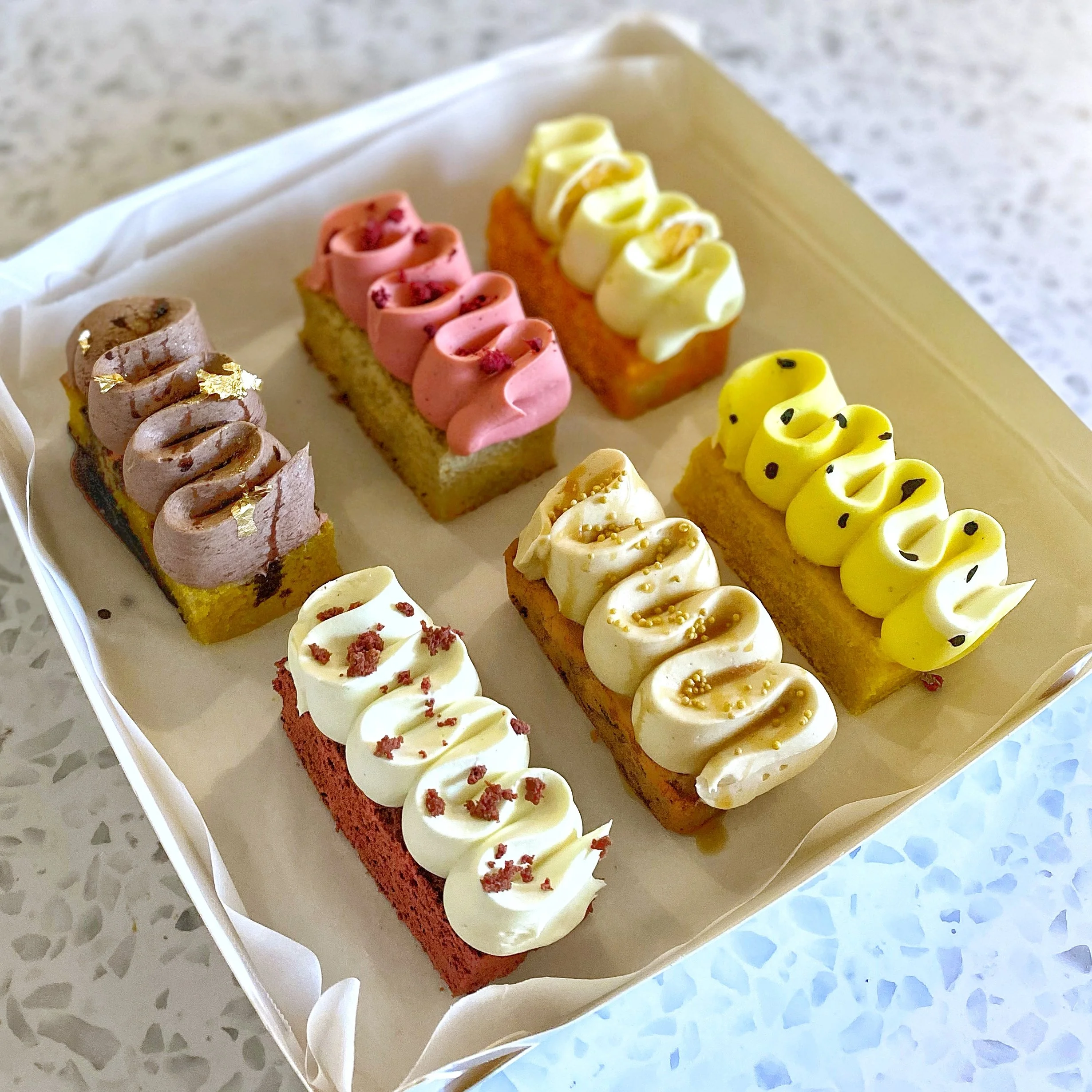 Six rectangular pieces of cake with colorful frosting and toppings arranged in a white tray.