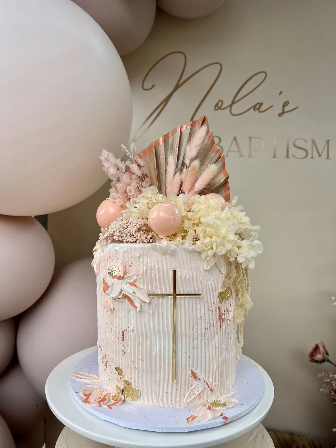 Pink birthday cake decorated with flowers, feathers, and gold accents, featuring a gold cross on the front, placed on a white cake stand with balloons and a backdrop reading "Nola's Baptism" in the background.