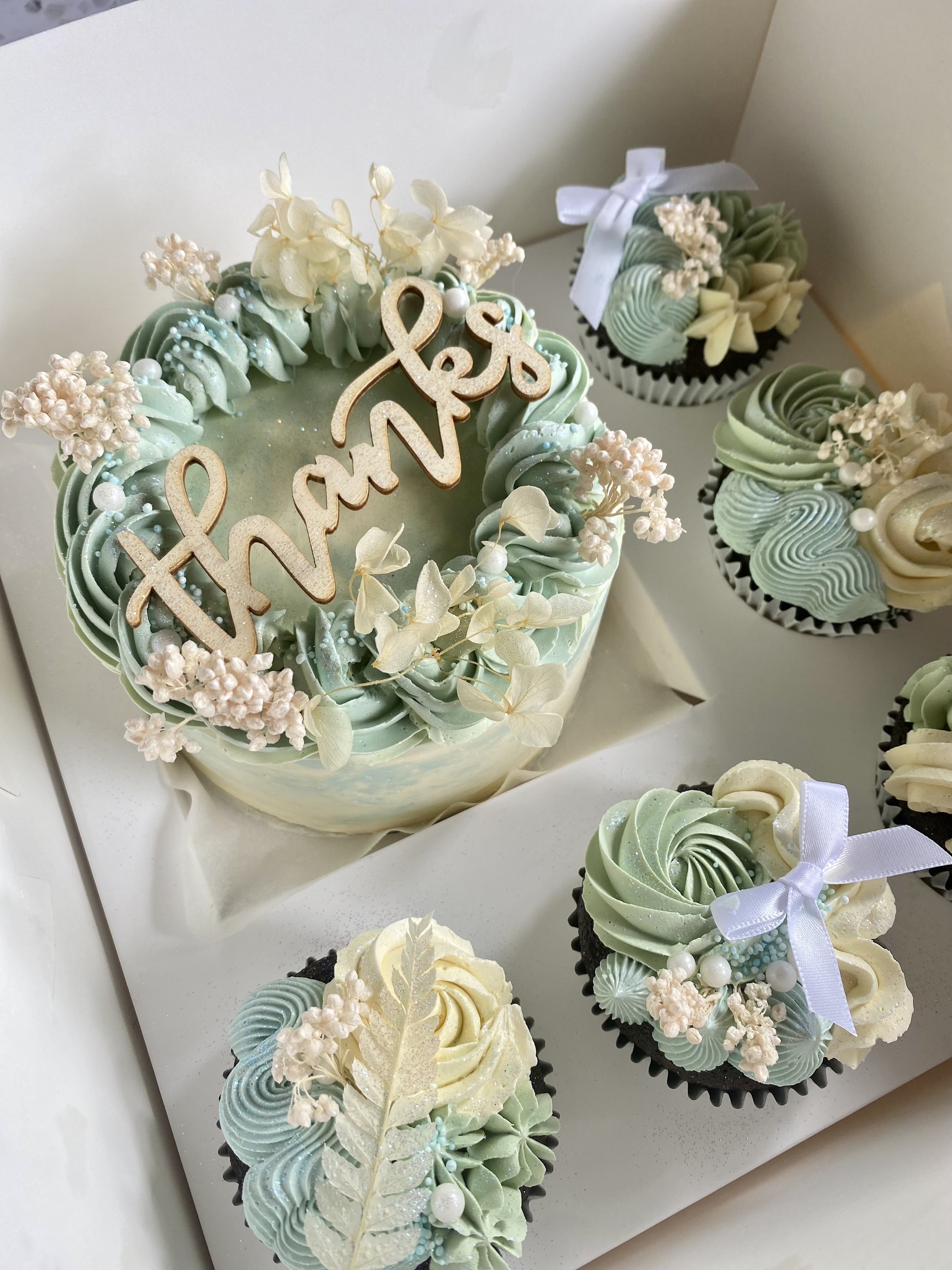 Cream-colored cake with pastel green and white frosting, floral decorations, and a "thanks" topper, surrounded by matching cupcakes with bows and floral accents.