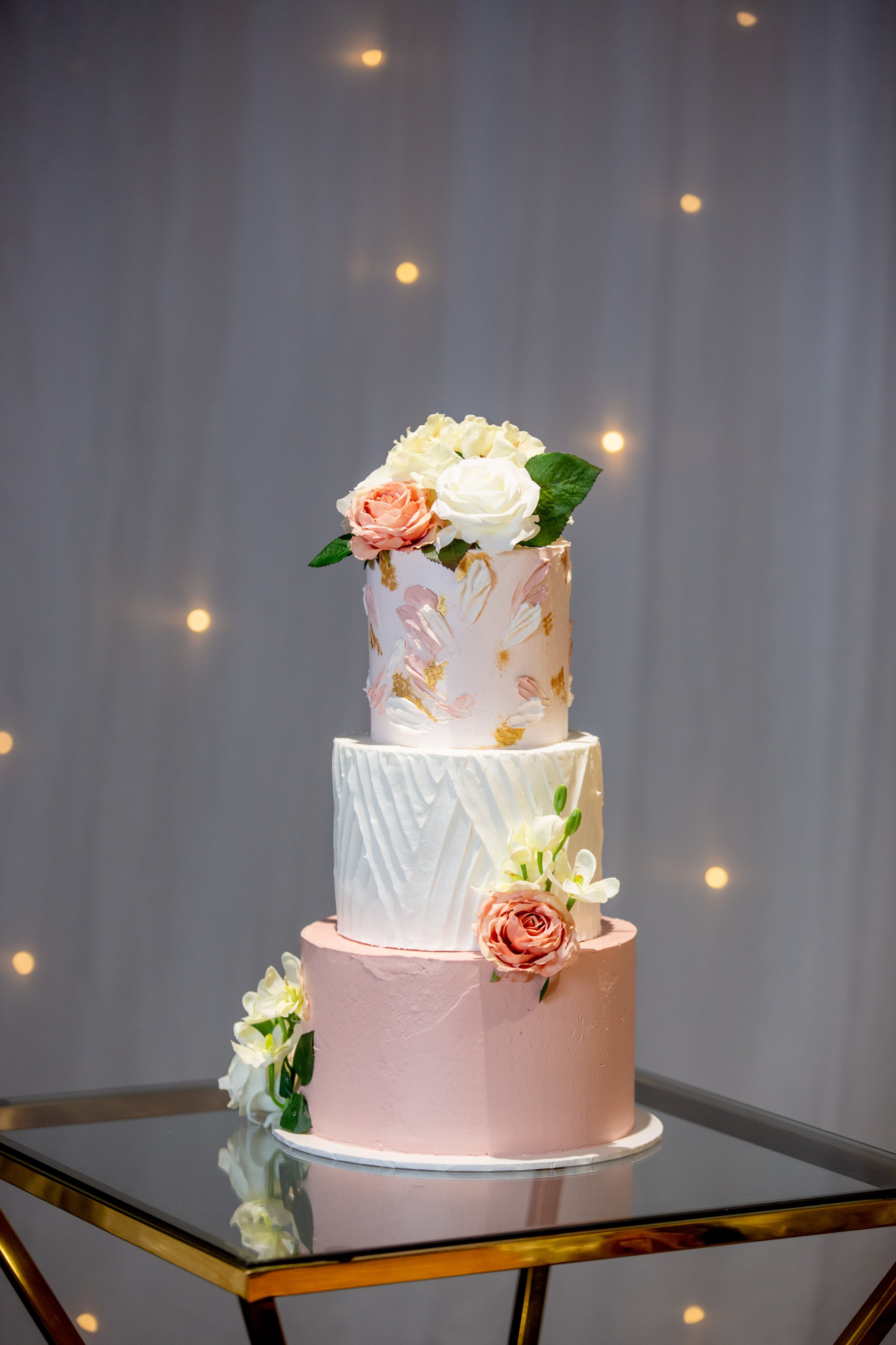 A three-tiered wedding cake with pink and white icing, decorated with pink and white roses and green leaves, placed on a reflective black surface against a gray background with small warm light spots.