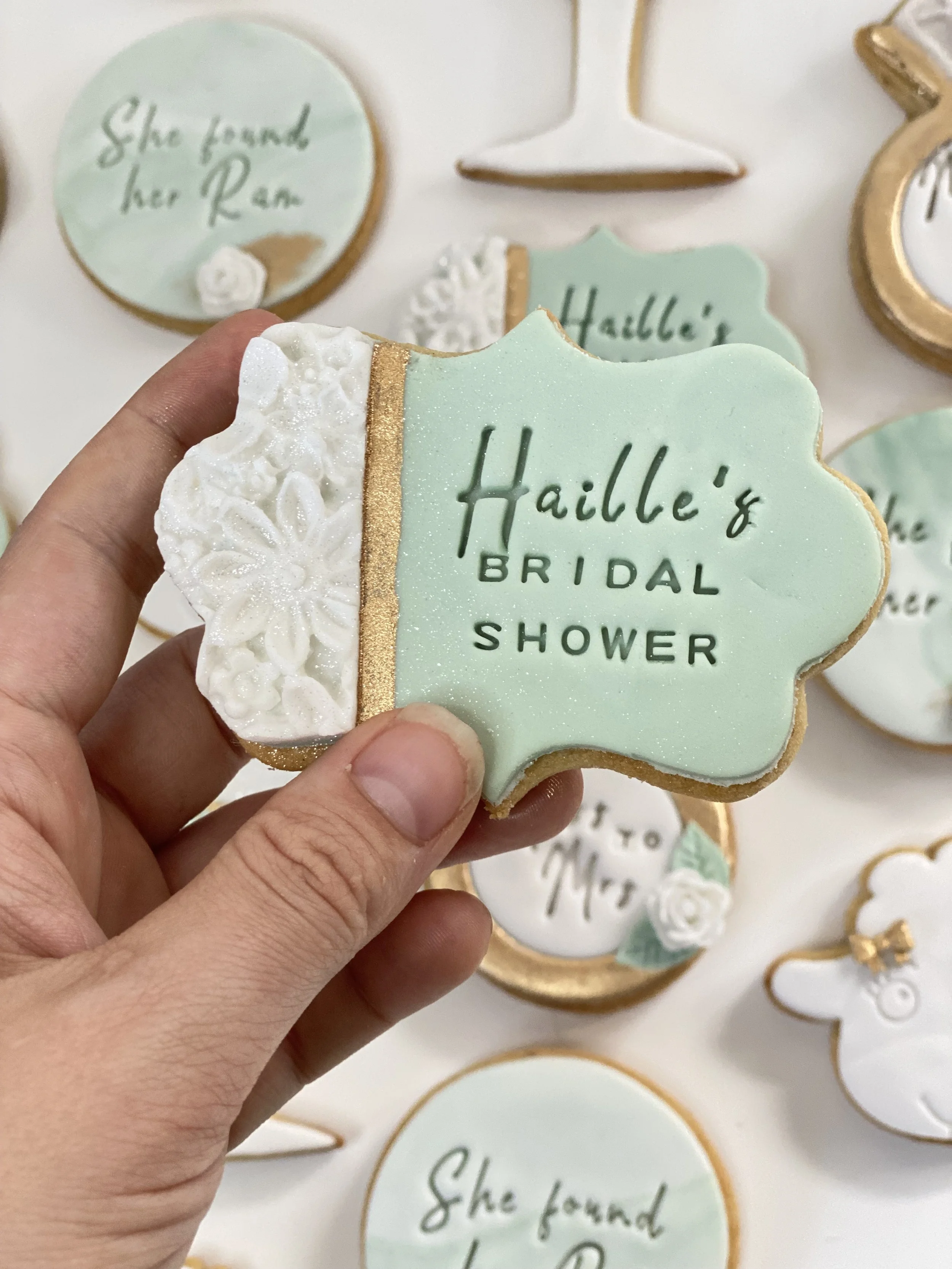 A decorated bridal shower cookie with light green icing, gold accents, and white floral icing, held in a person's hand. Other similar cookies are visible in the background.