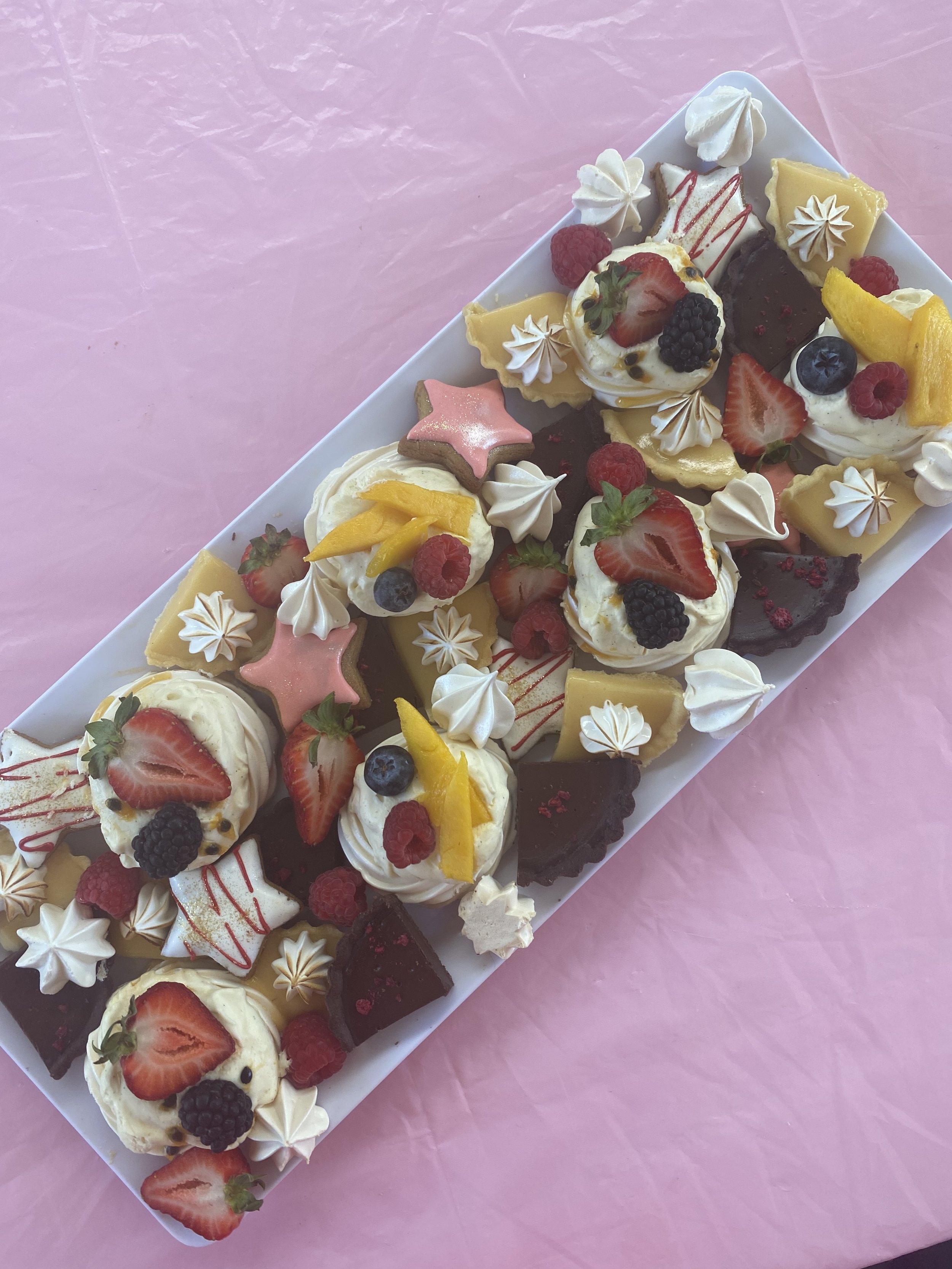 Assorted mini desserts with fresh berries, fruit, and decorative icing on a white platter against pink background.