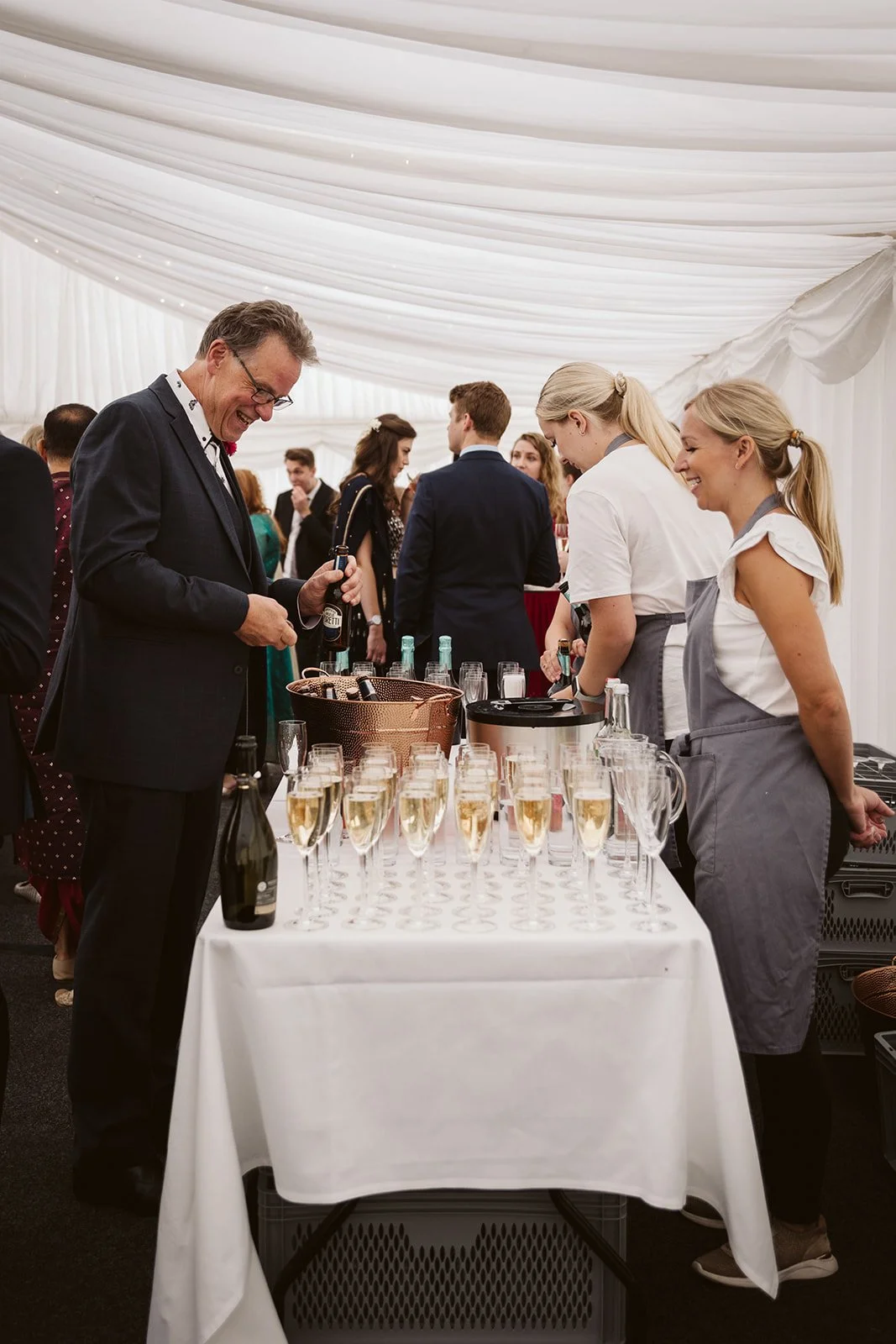 People enjoying a drinks bar at a social event, with champagne flutes filled with a bubbly beverage on a white tablecloth, inside a white tent.