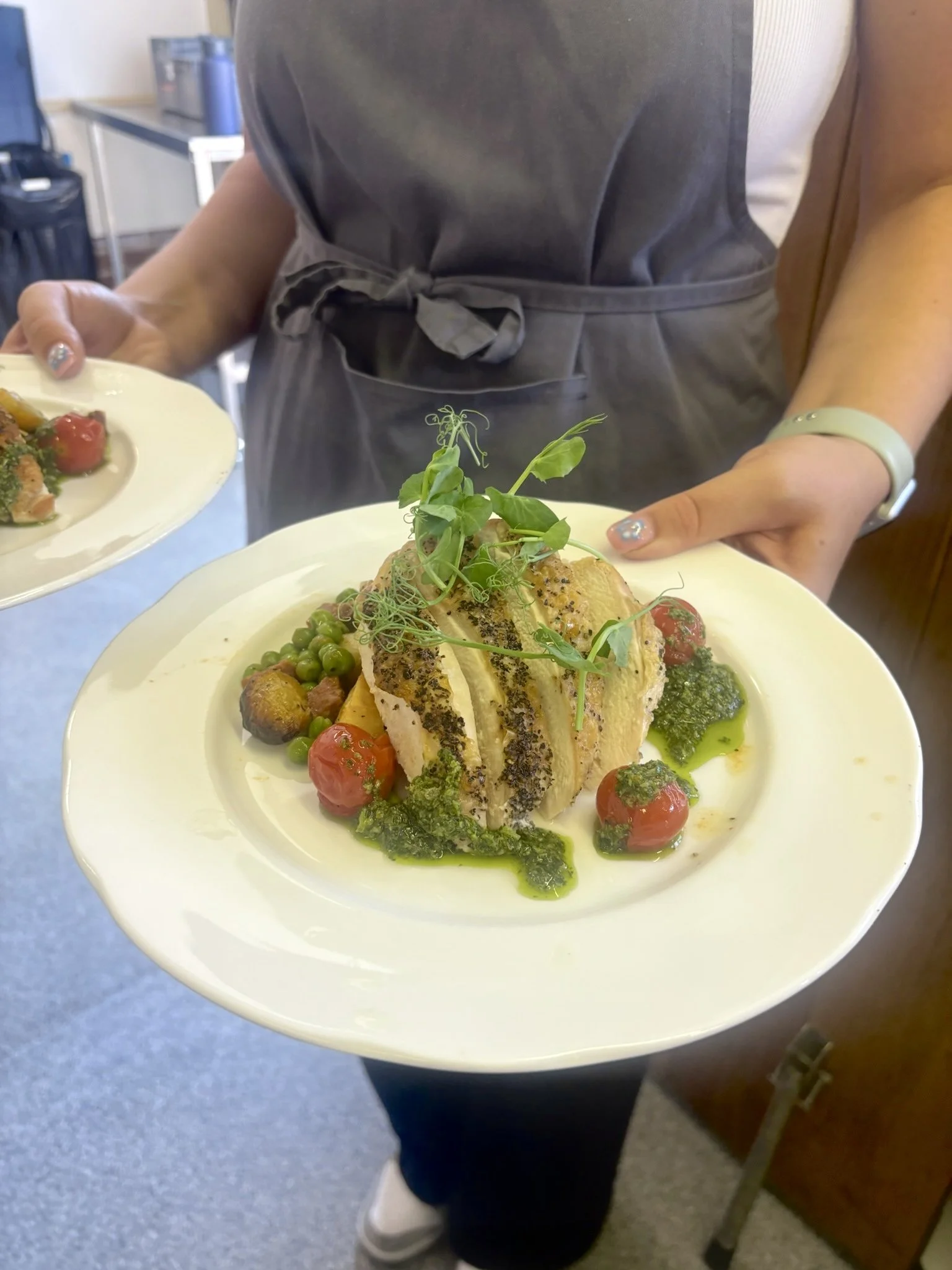 Person holding a white plate with a plated dish of stuffed chicken breast topped with microgreens, surrounded by cherry tomatoes, green peas, broccoli, and green sauce.