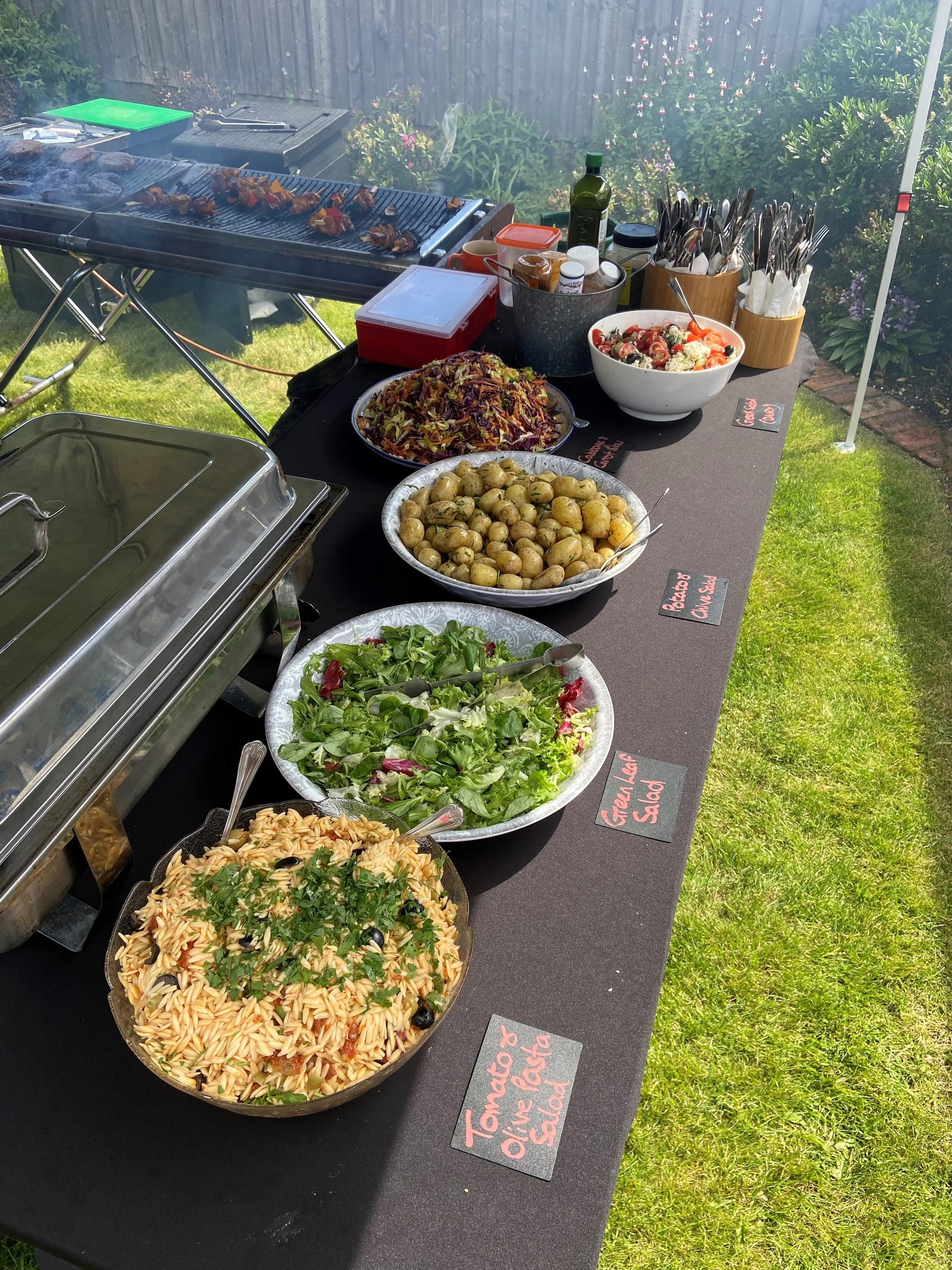 Outdoor buffet table with various salads, roasted potatoes, and a bowl of caprese salad, with a grill in the background cooking skewered meat and vegetables.