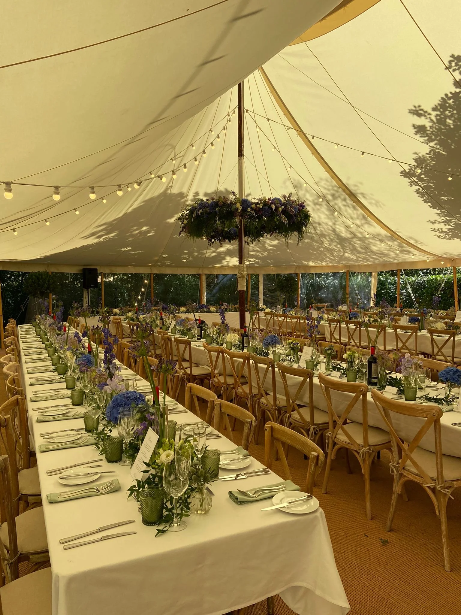 Underground event tent decorated with hanging string lights and floral arrangements, with a long banquet table set with flowers, glasses, and utensils for a celebration.