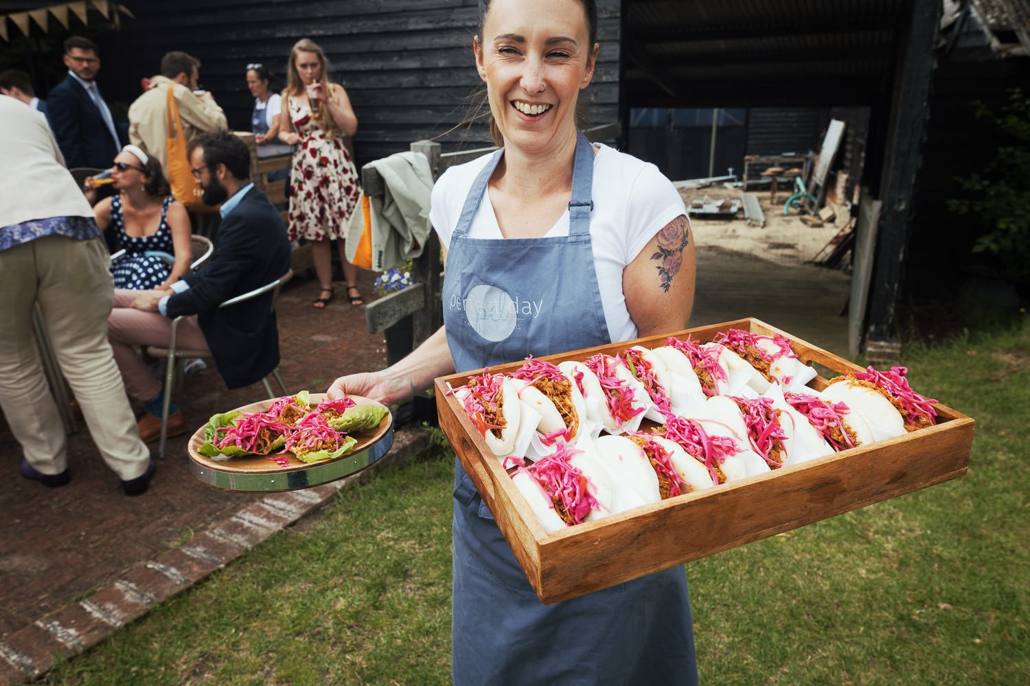 A smiling woman wearing an apron holding a tray of tacos topped with pickled red onions, with a group of people in the background at an outdoor gathering.