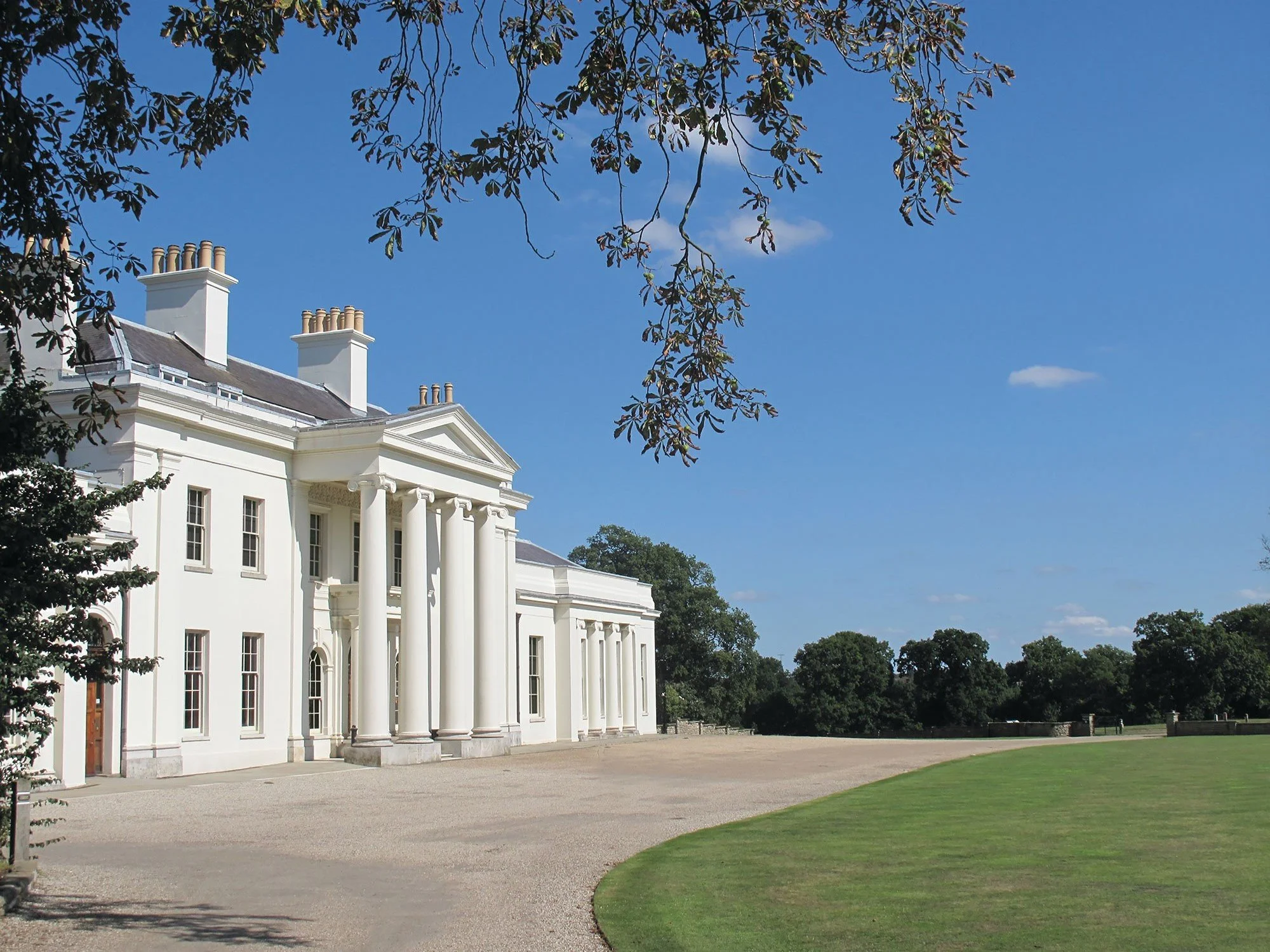 White mansion with Greek columns and a main entry door, surrounded by green lawn and trees, under a clear blue sky.