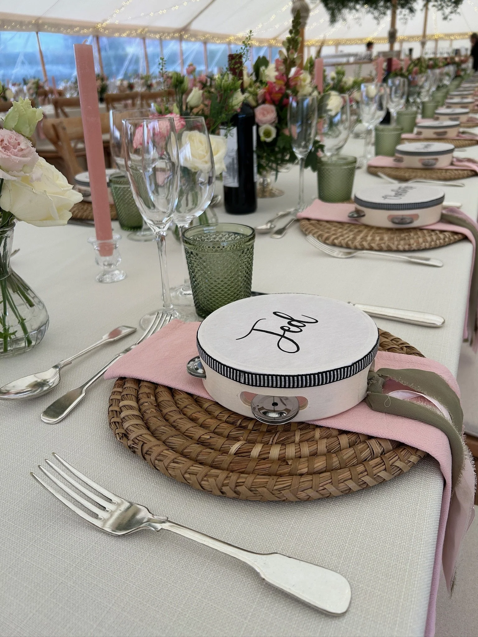 Set up for a wedding reception with a long table decorated with pink, white, and green floral centerpieces, pink candles, and glassware. Each place setting has woven placemats, pink napkins, a small pink box labeled "food," and wedding cake servers.