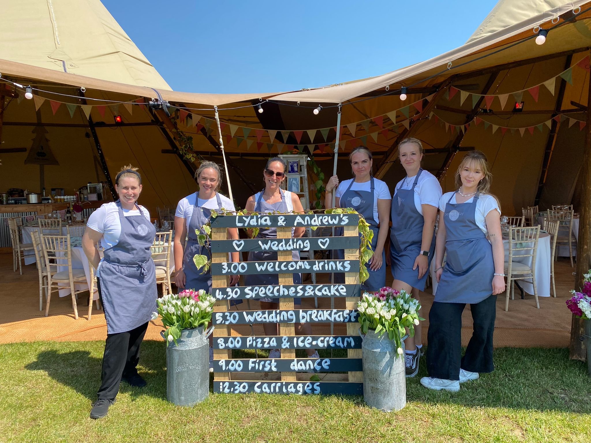 Group of six women wearing aprons standing behind a wedding schedule sign outside a large tent decorated with bunting and string lights.