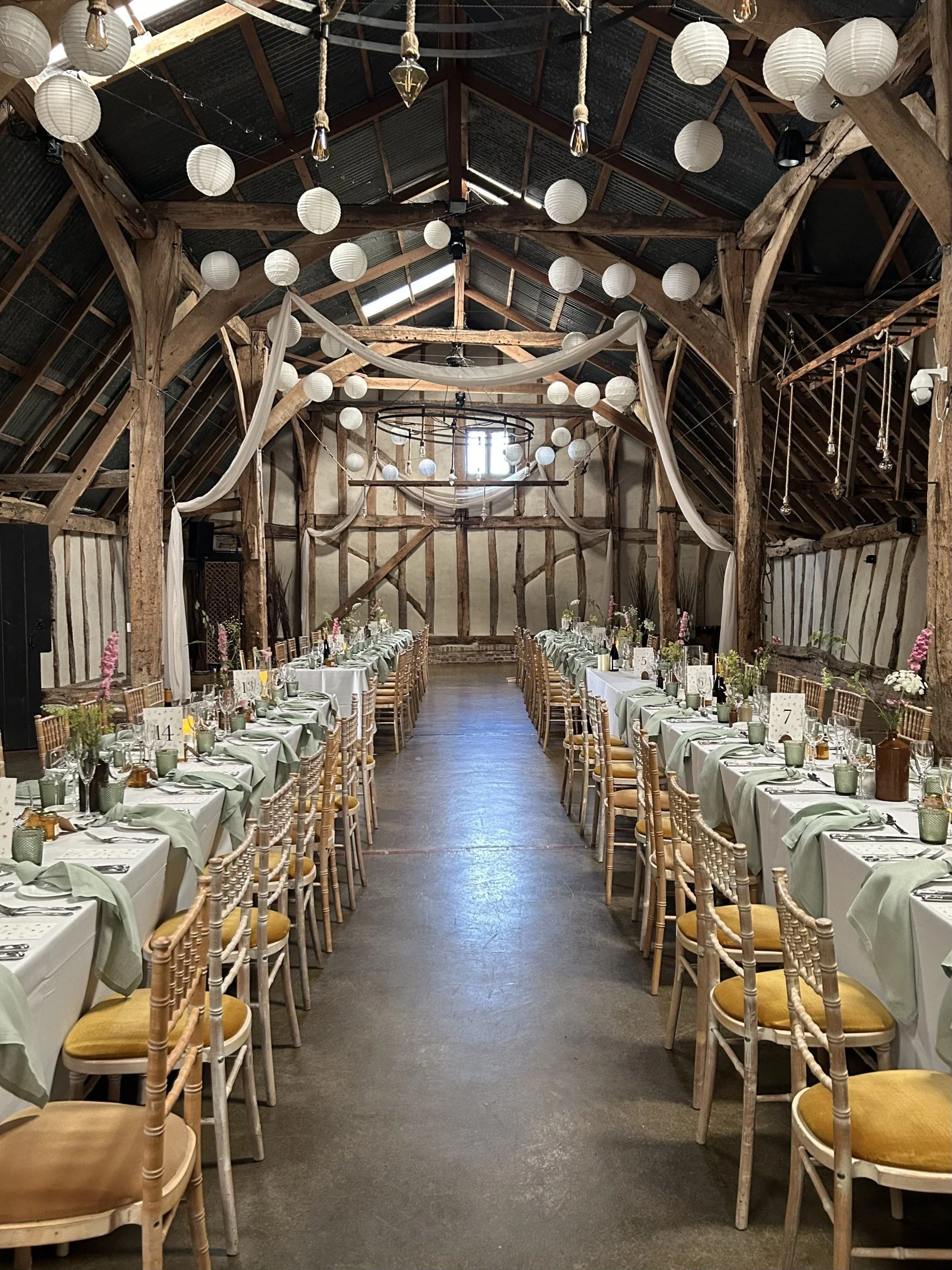 Wedding reception setup inside a rustic barn with long tables, decorated with white tablecloths, vases of flowers, candles, and table number cards. White paper lanterns and draped fabric hang from the ceiling.
