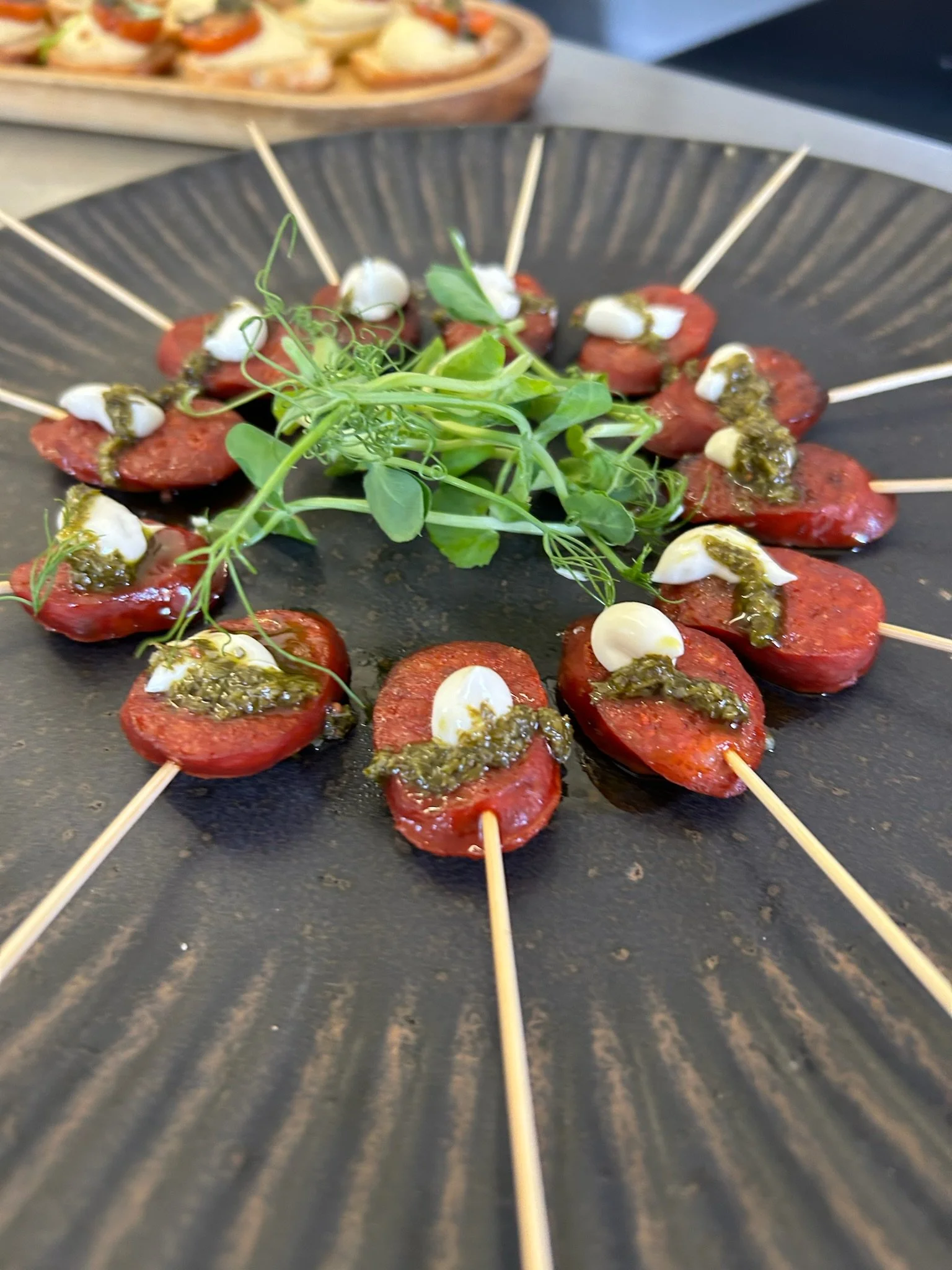 Skewered cherry tomatoes topped with dollops of creamy sauce and green pesto, garnished with fresh microgreens on a black plate.