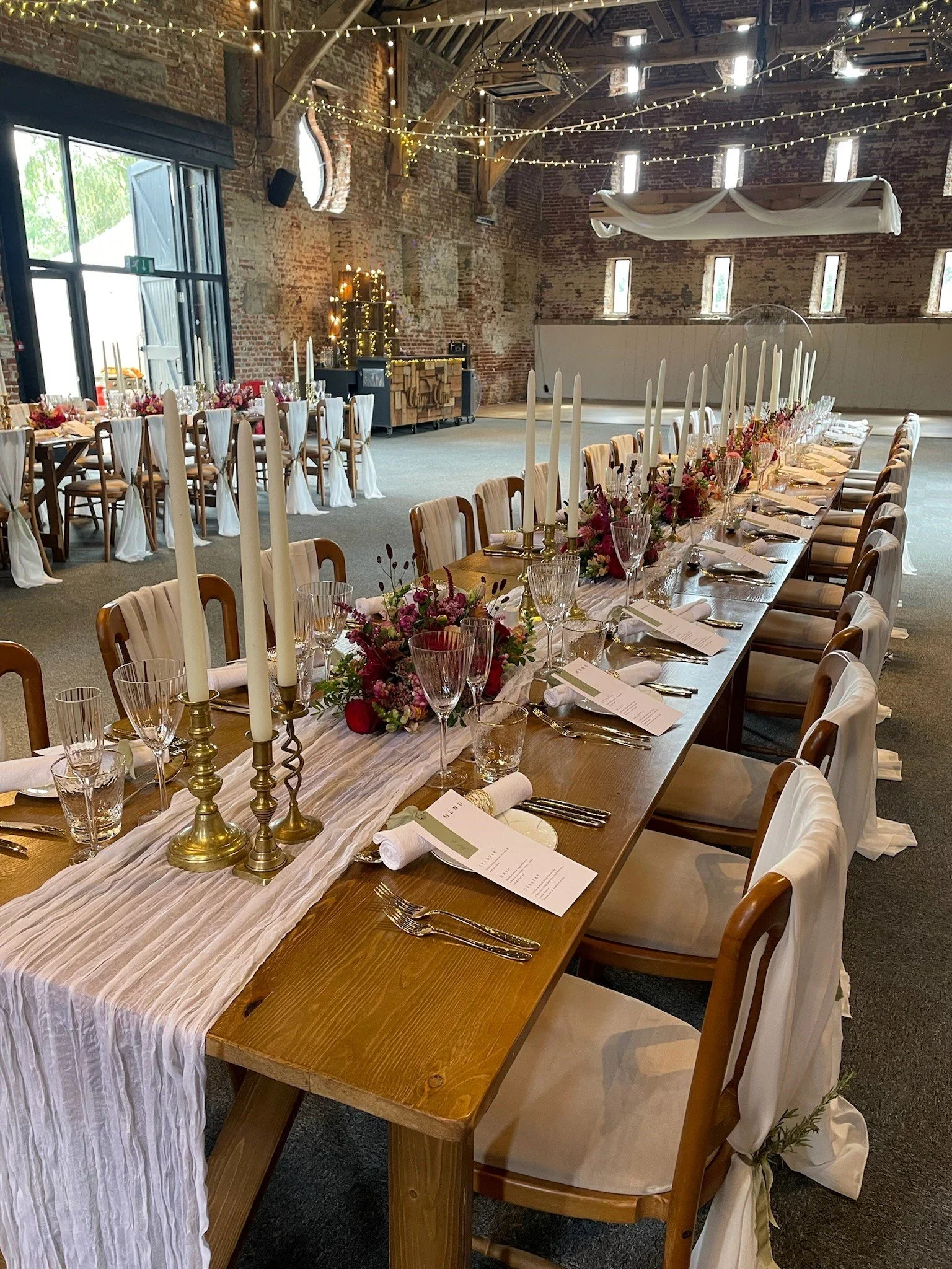 Long wooden dining table set up for a wedding reception with white chair covers, floral centerpieces, tall white candles, menus, and glassware, inside a rustic brick venue decorated with string lights.