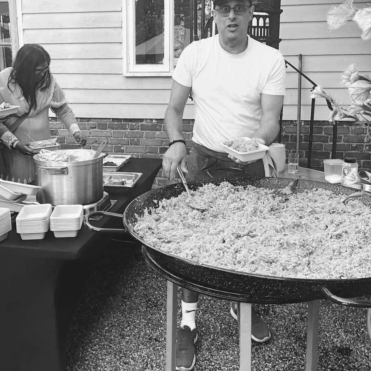 A man serving food from a large pan at an outdoor event, with a woman in the background preparing additional dishes.
