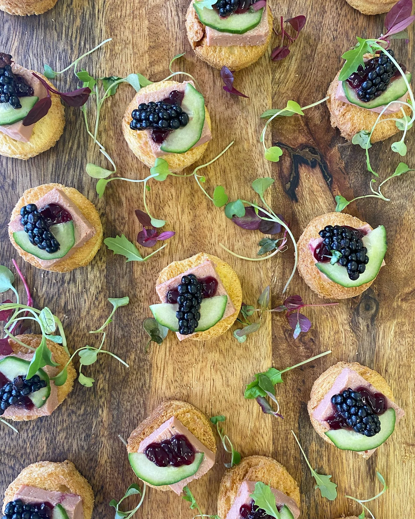 Assorted canapé bites on a wooden board, topped with cucumber slices, blackberries, and a spread, decorated with microgreens.