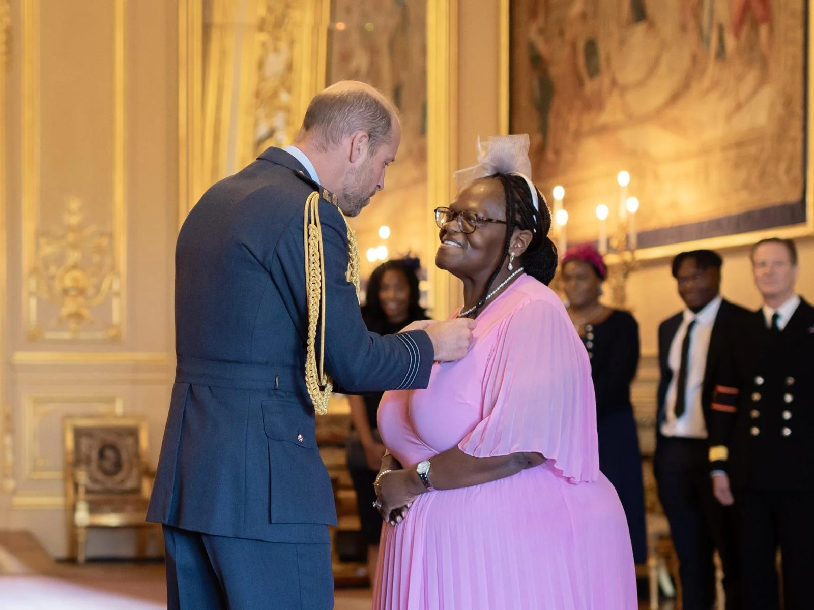 A man in a military uniform is pinning a medal on a woman wearing glasses and a pink dress in an ornate room with gold accents and chandeliers, with several people in formal attire observing.