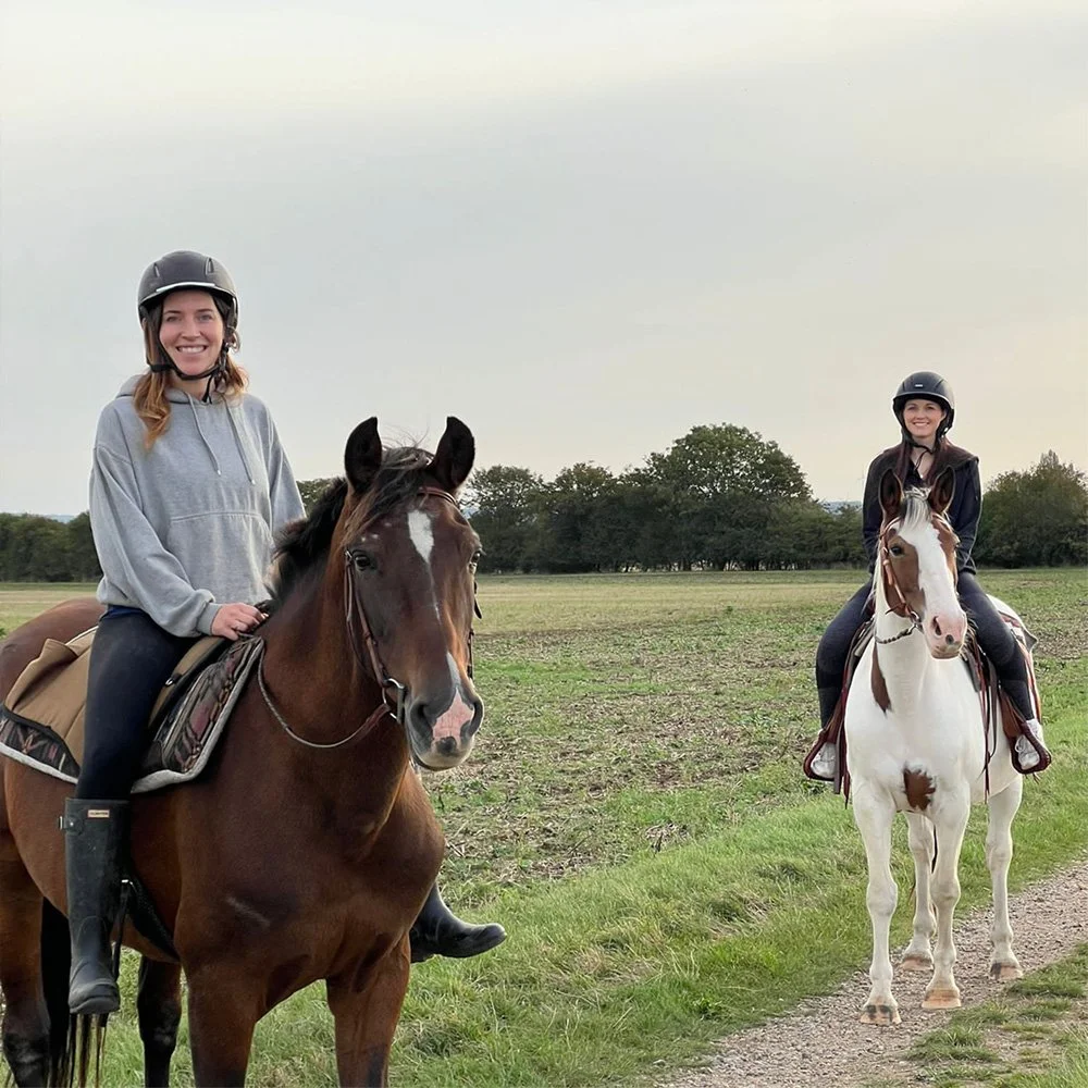 Two women riding horses in an open field, both wearing riding helmets and smiling.