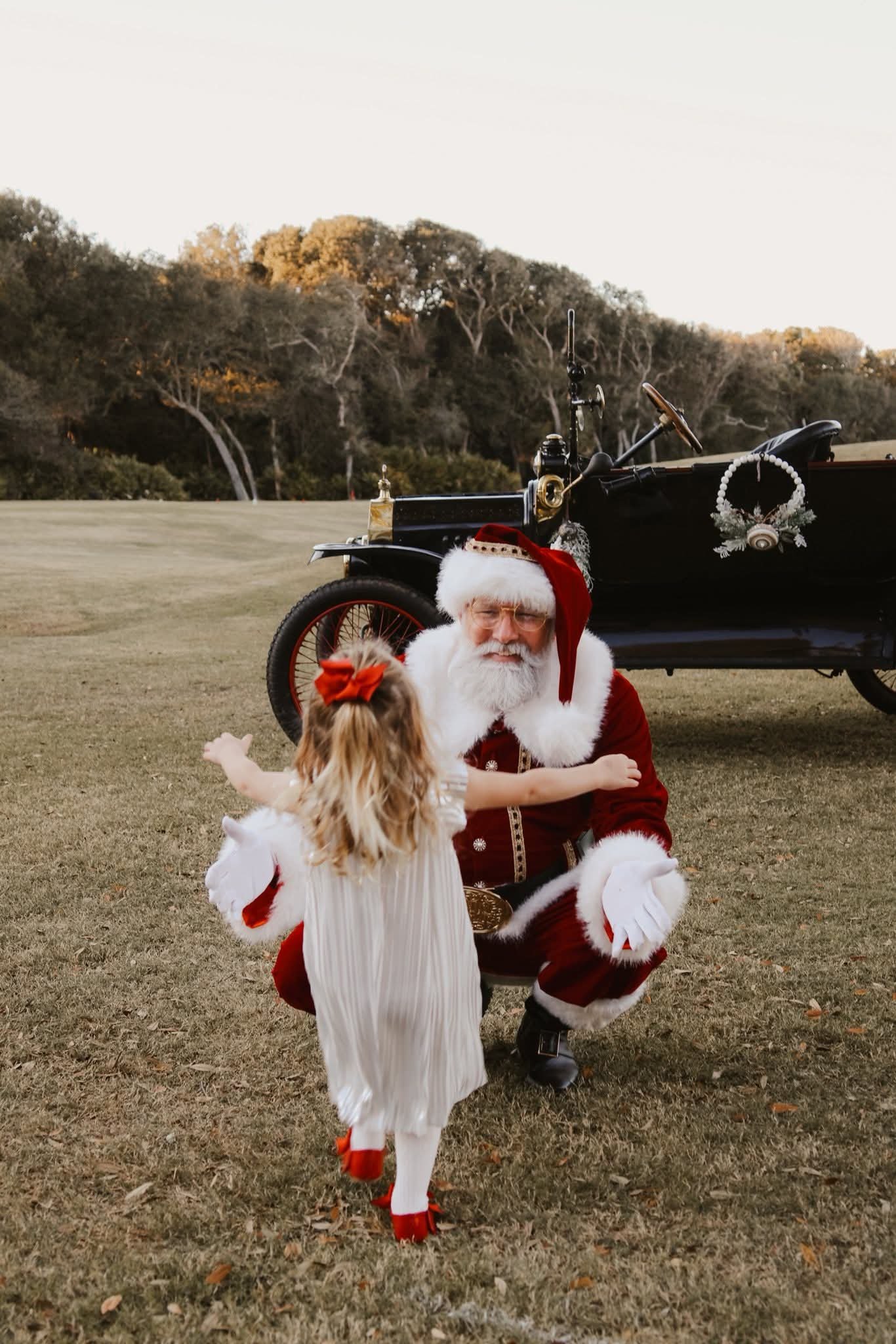Santa Claus kneeling on the grass, embracing a young girl. Santa is in a traditional red and white outfit with a fur-trimmed hat, white gloves, and a white beard. The girl, with blonde hair and a red bow, is reaching out to Santa. Behind them is a vintage black car decorated with Christmas ornaments, in an open outdoor field with trees in the background.
