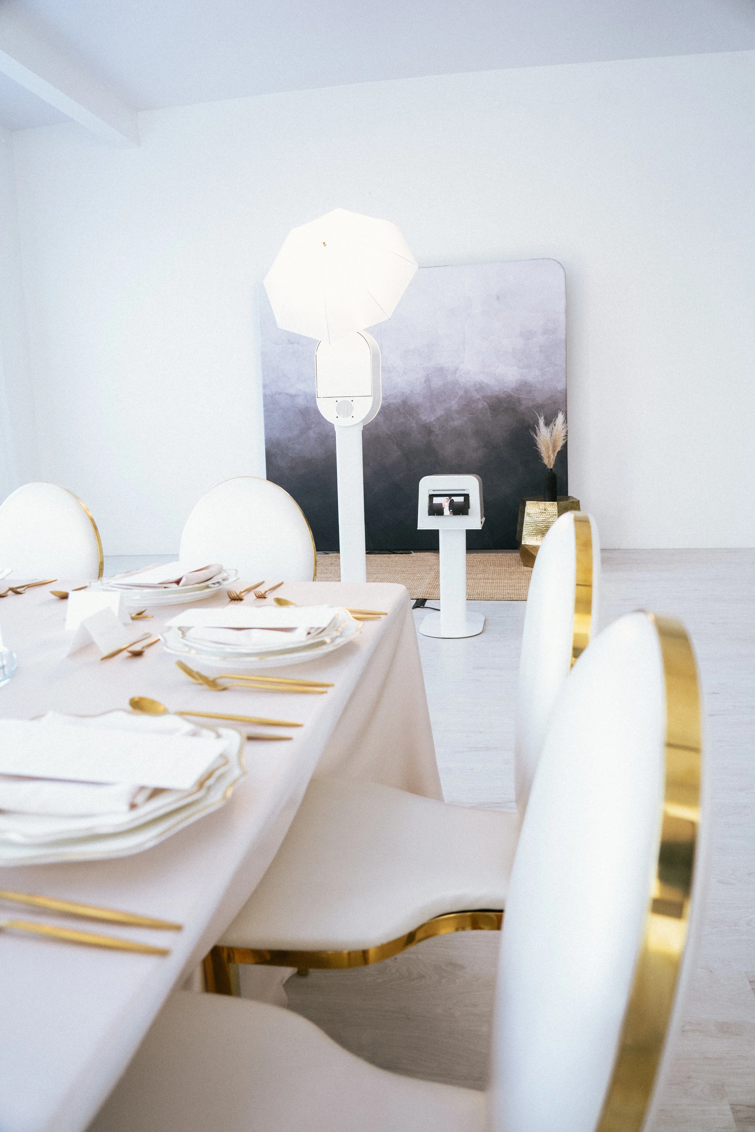 Modern indoor space with white chairs featuring gold accents, a dining table set with white plates and gold utensils, a large photography backdrop, and a ring light with umbrella, indicating a photo booth area.
