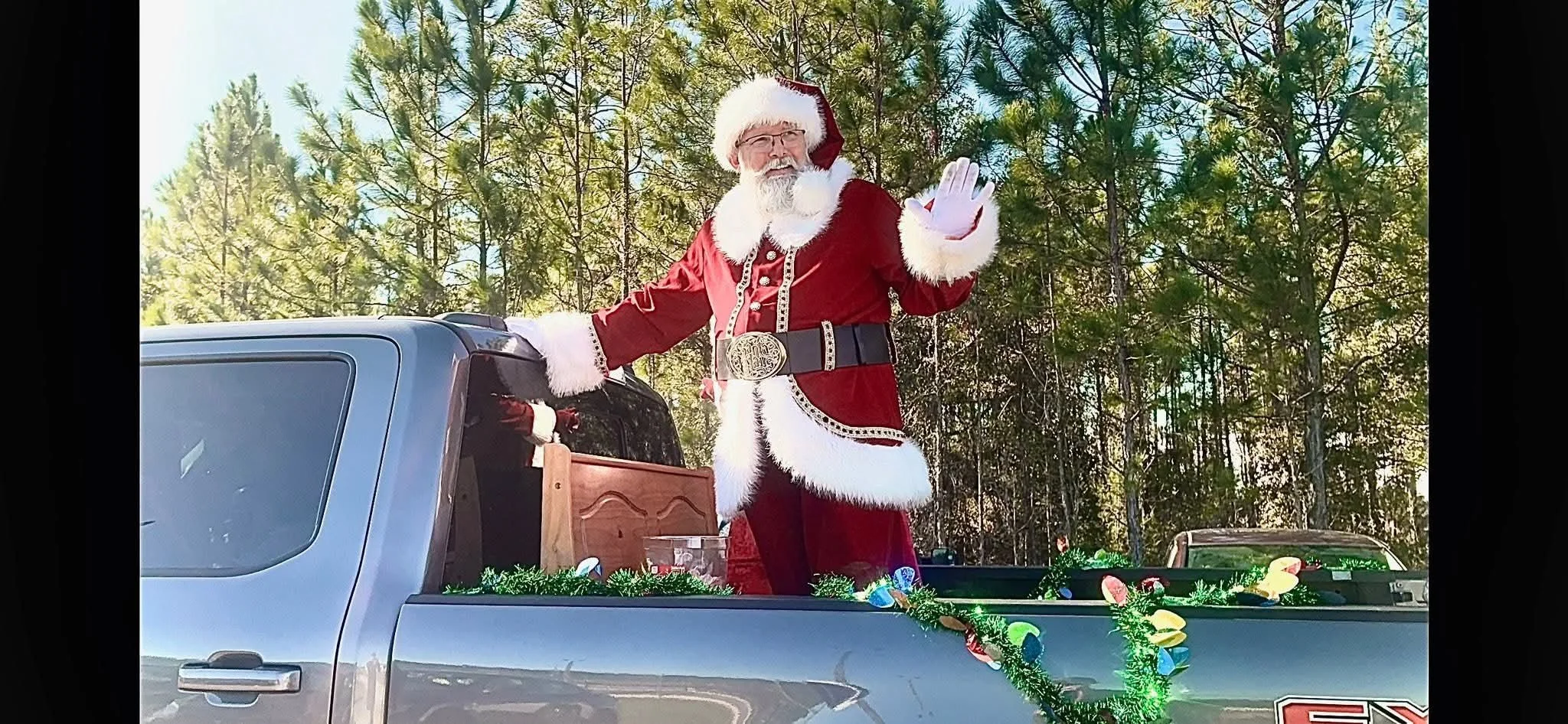 Santa Claus dressed in a red suit with white trim, waving from the back of a decorated truck.