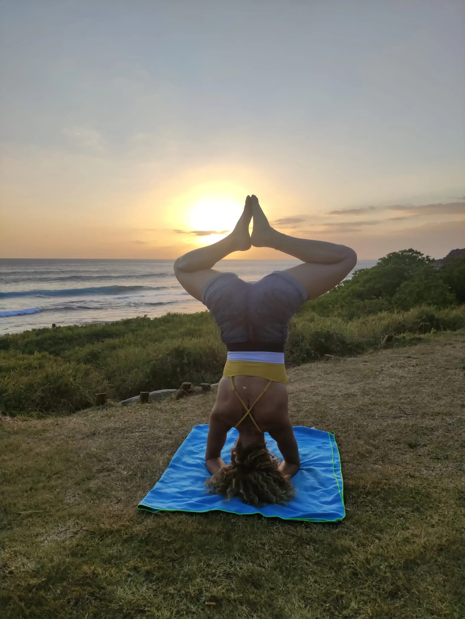 Une femme faisant de la pose de yoga en équilibre sur sa tête sur un tapis en bord de mer au coucher du soleil.