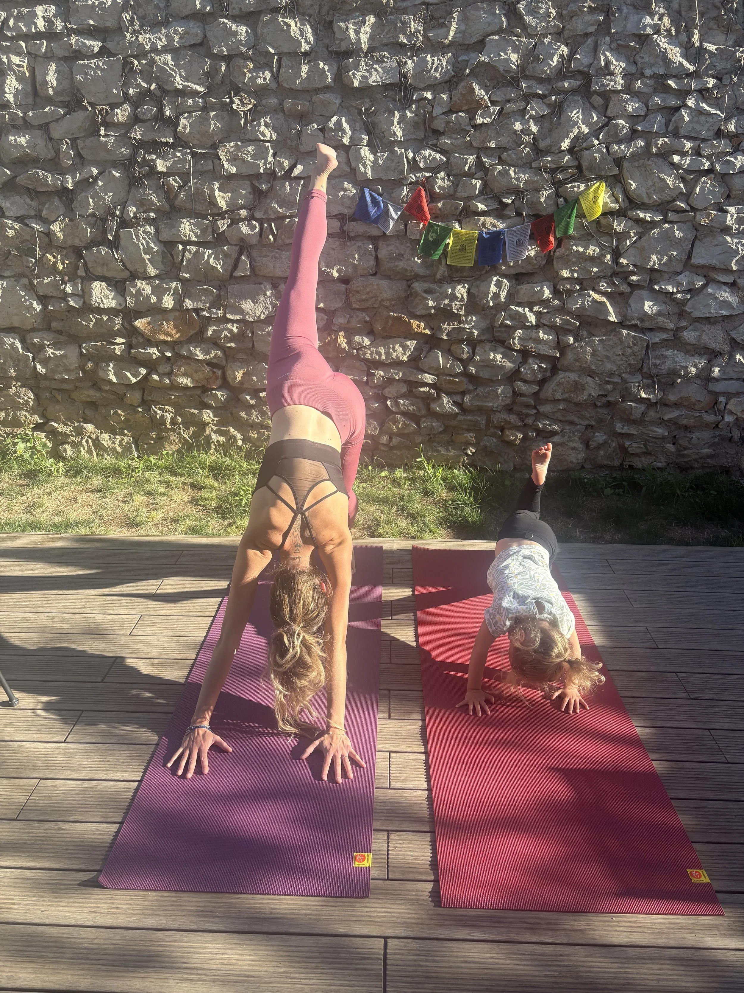 Deux personnes, une adulte et une enfant, pratiquent le yoga en position de planche inversée sur des tapis de yoga roses et rouges en plein air, face à un mur de pierre avec des drapeaux colorés suspendus.