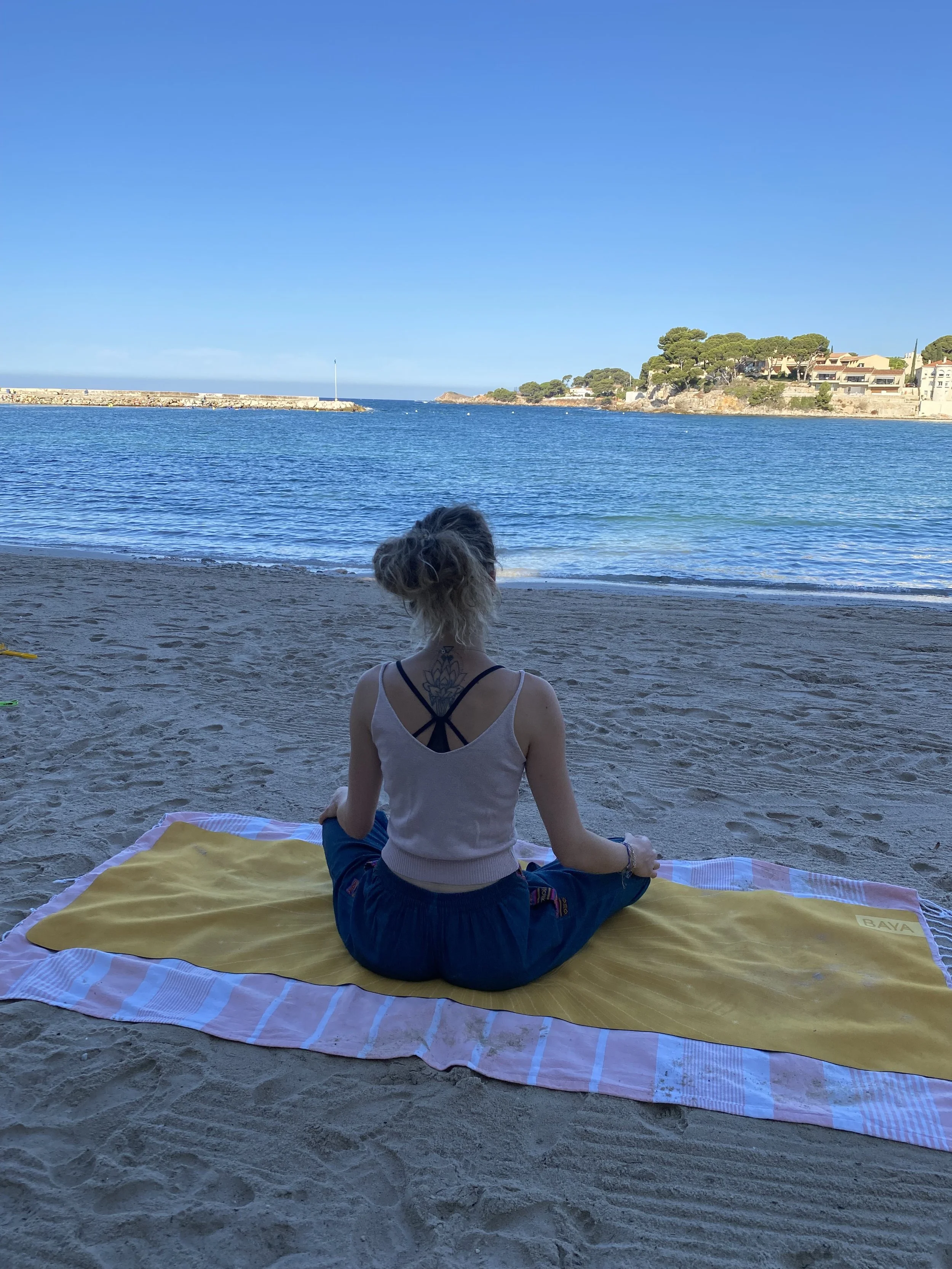 Une femme assise en tailleur sur une serviette jaune sur une plage, regardant la mer, avec des maisons et des arbres en arrière-plan sur la côte sous un ciel clair.