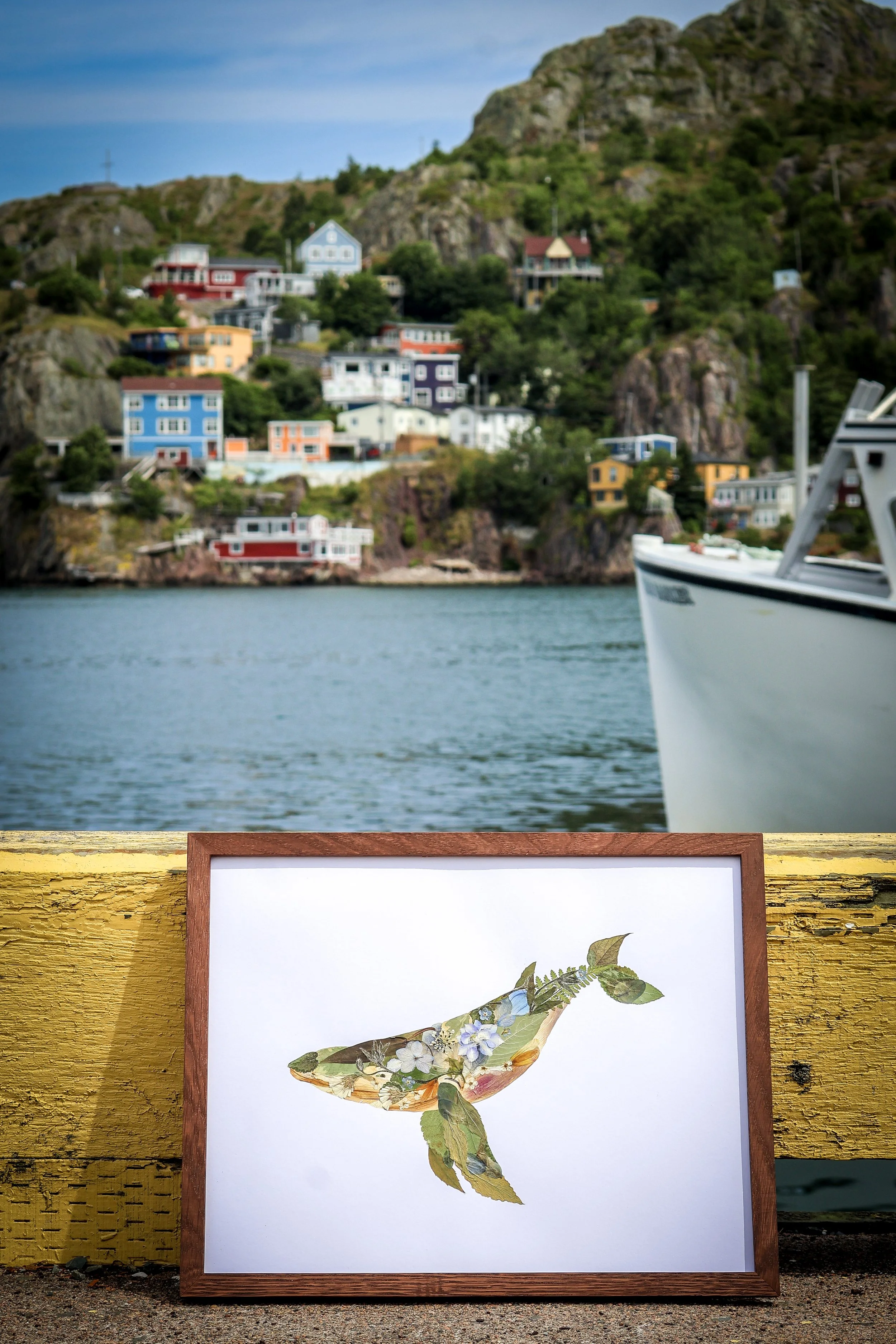 A framed artwork of a whale made from botanical illustrations, resting on a yellow wooden railing near water, with colorful houses on a hillside and a boat in the water.