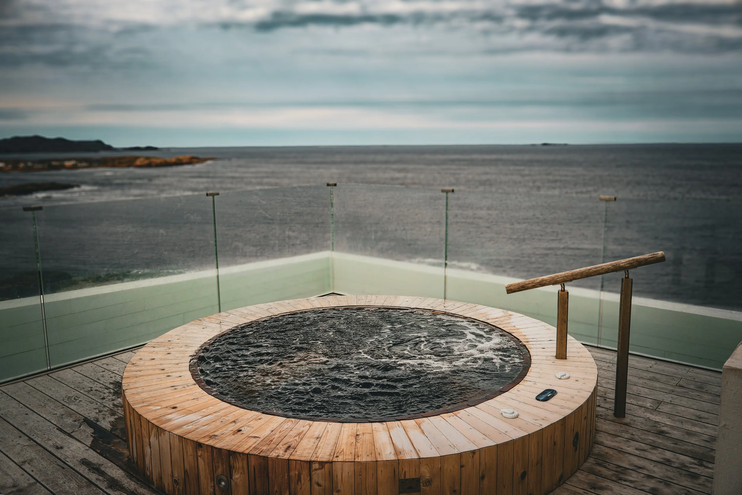 Hot tub on a wooden deck overlooking the ocean with a glass barrier and cloudy sky.