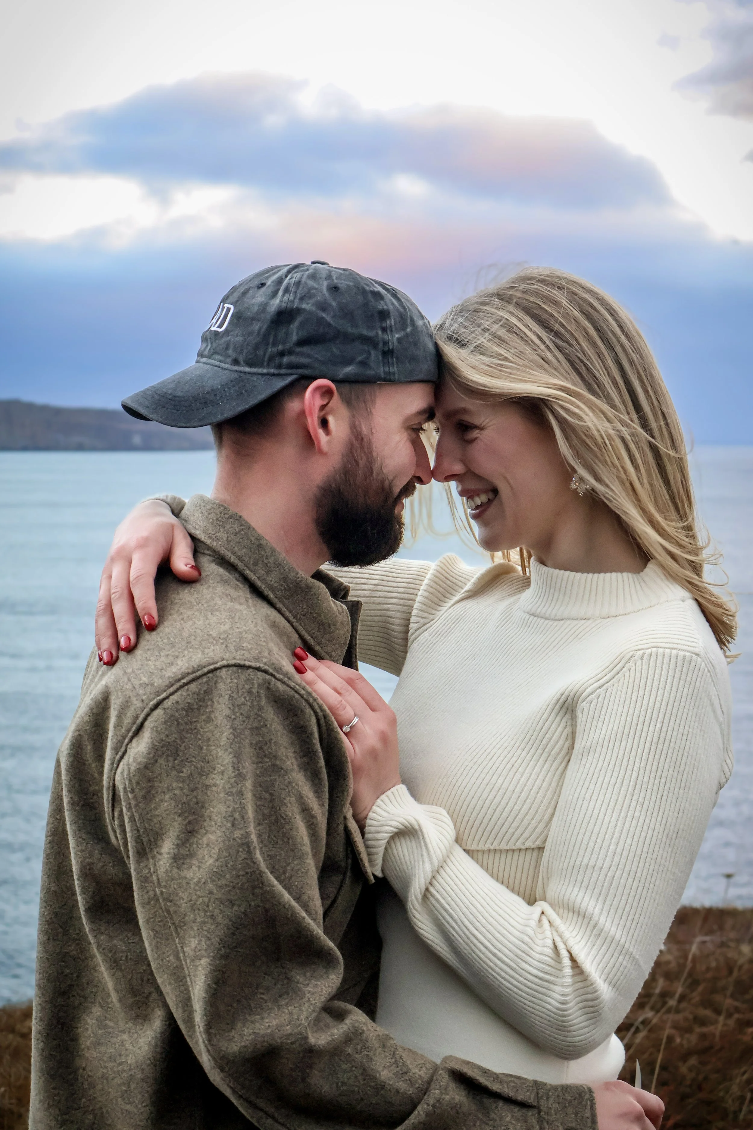 A couple embraces near a body of water with cliffs in the background during sunset, touching foreheads and smiling.