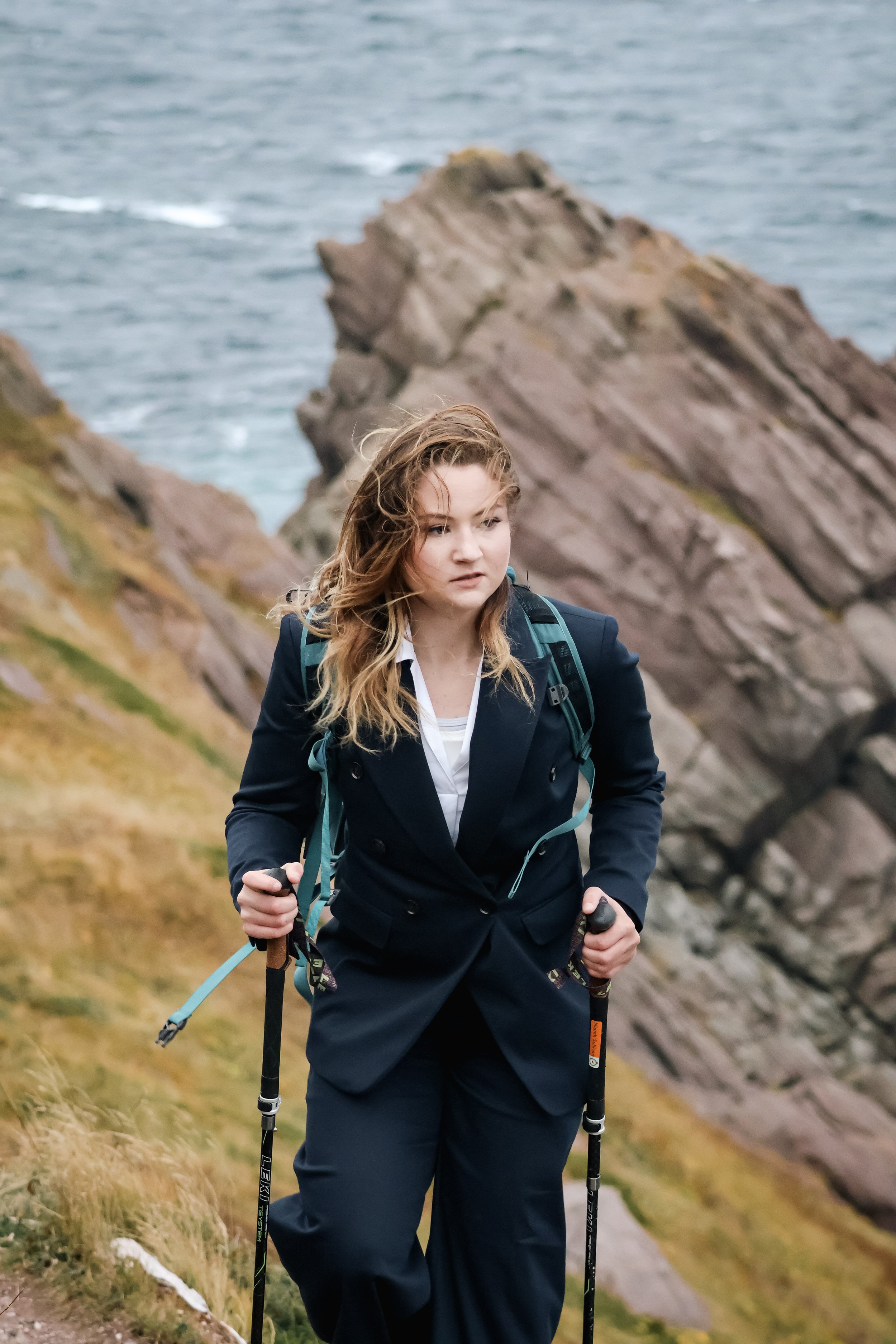 A woman hiking along a rocky coastal trail with a backpack and trekking poles, overlooking the ocean and rugged cliffs.