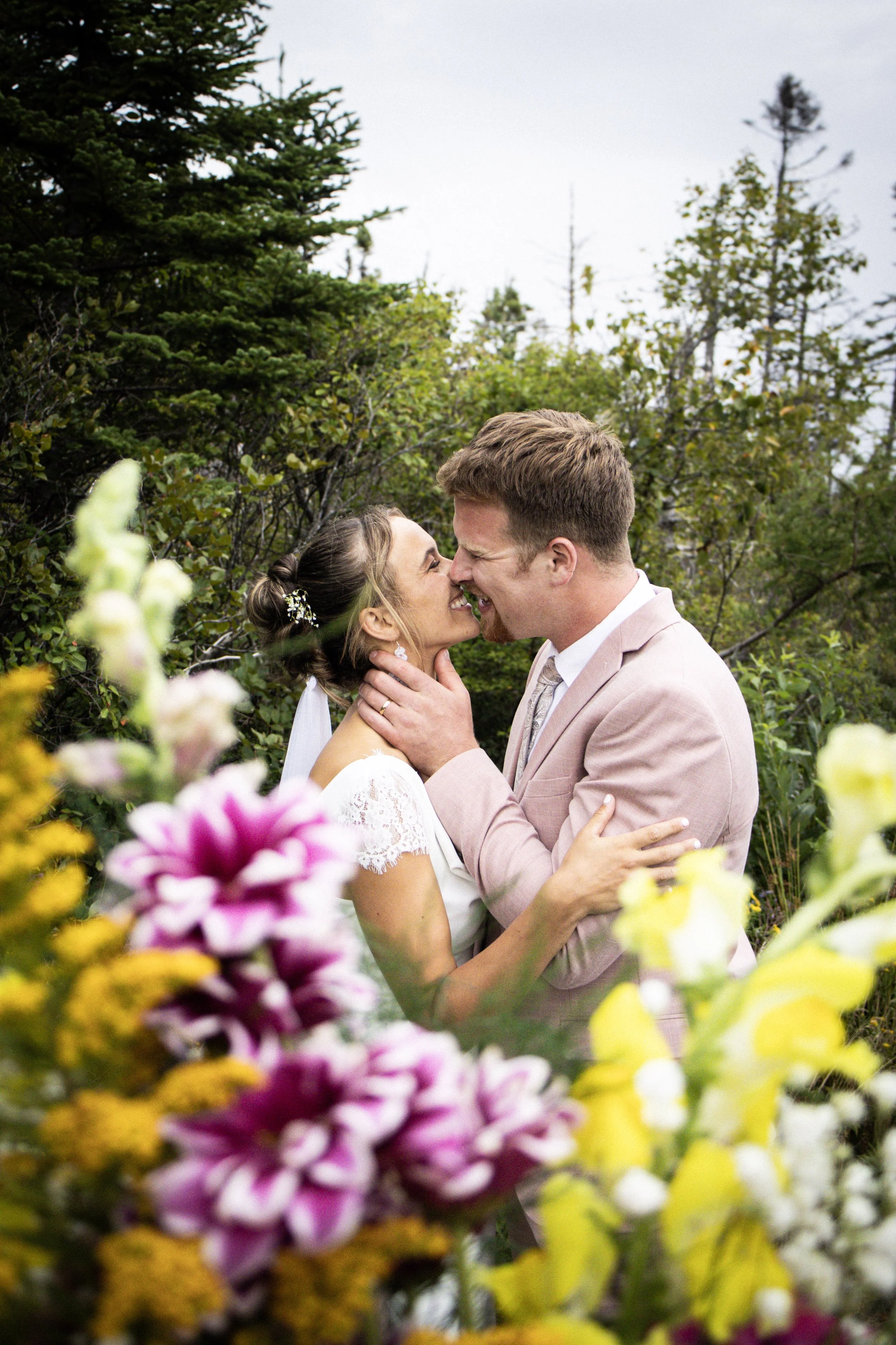 A bride and groom share a kiss in a garden, surrounded by colorful flowers and greenery.
