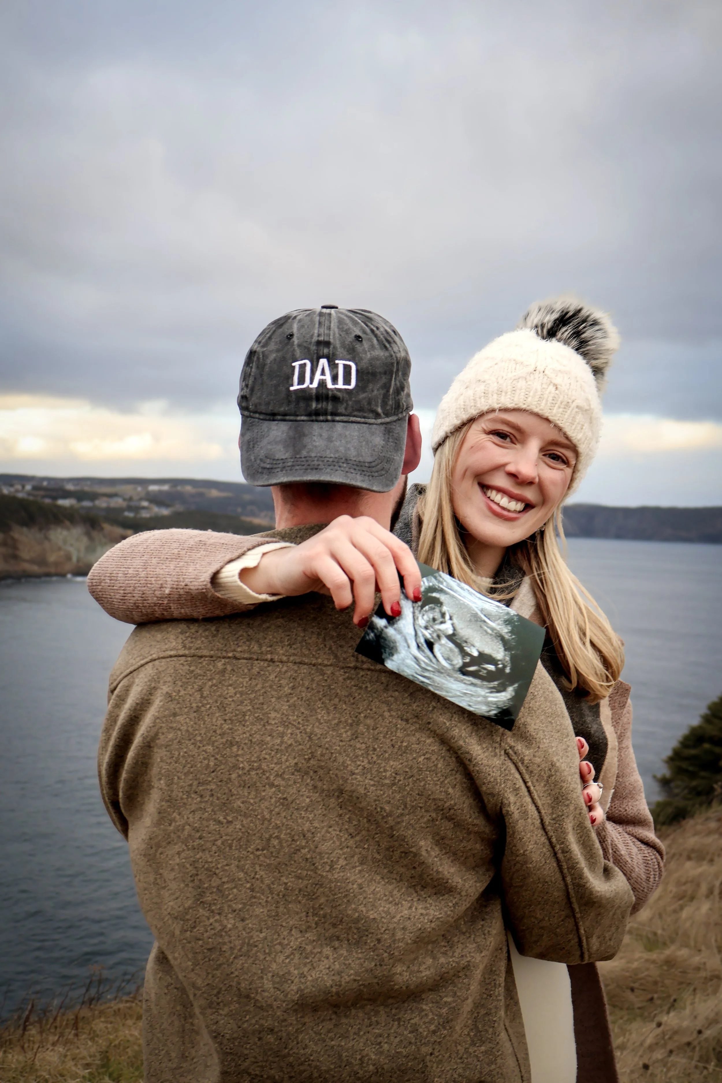 A woman smiling and holding an ultrasound picture, hugging a man wearing a cap that says 'DAD', standing outdoors near a body of water with cloudy skies.