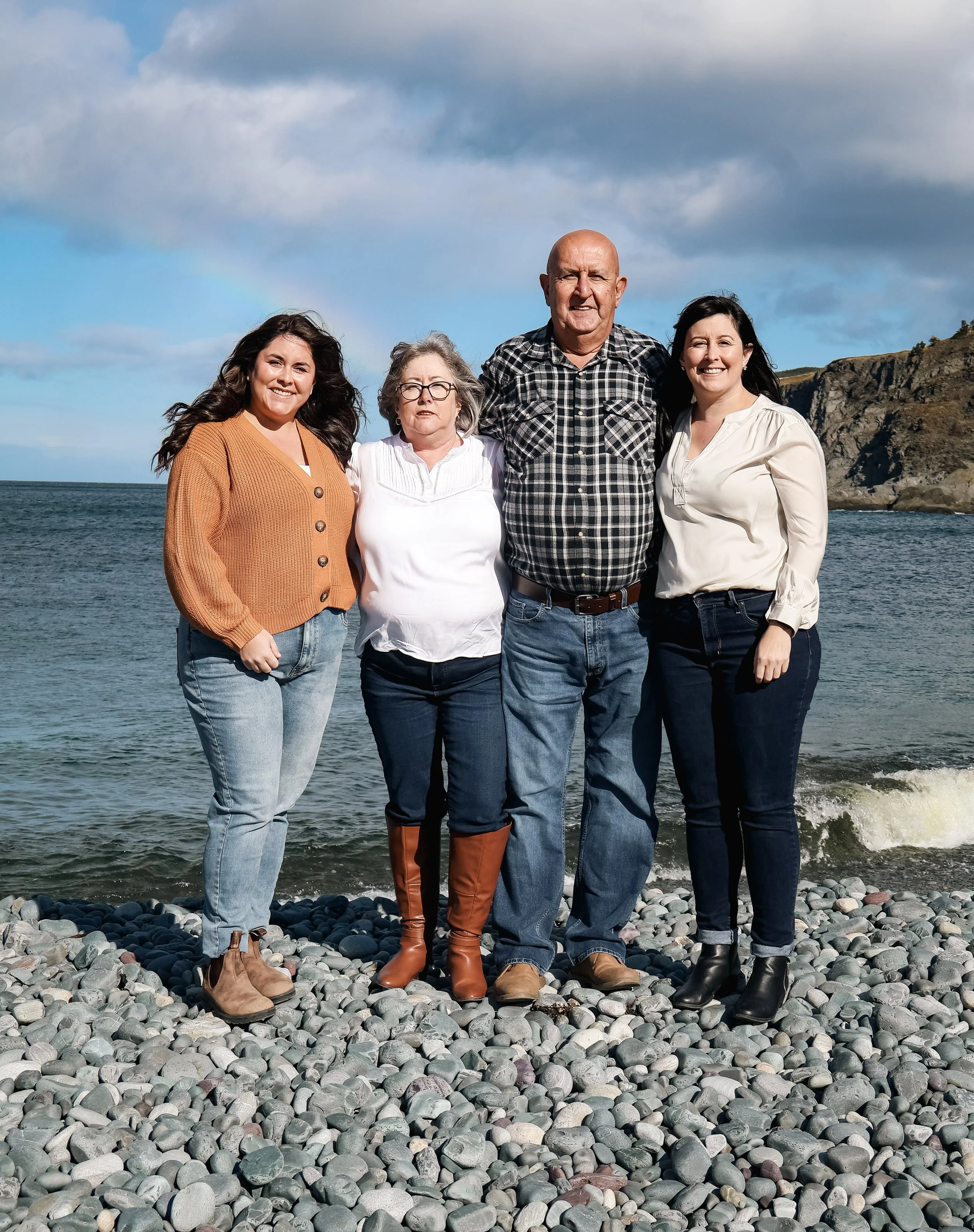 Family of five standing on rocky beach near ocean, with cliffs in the background and partly cloudy sky.