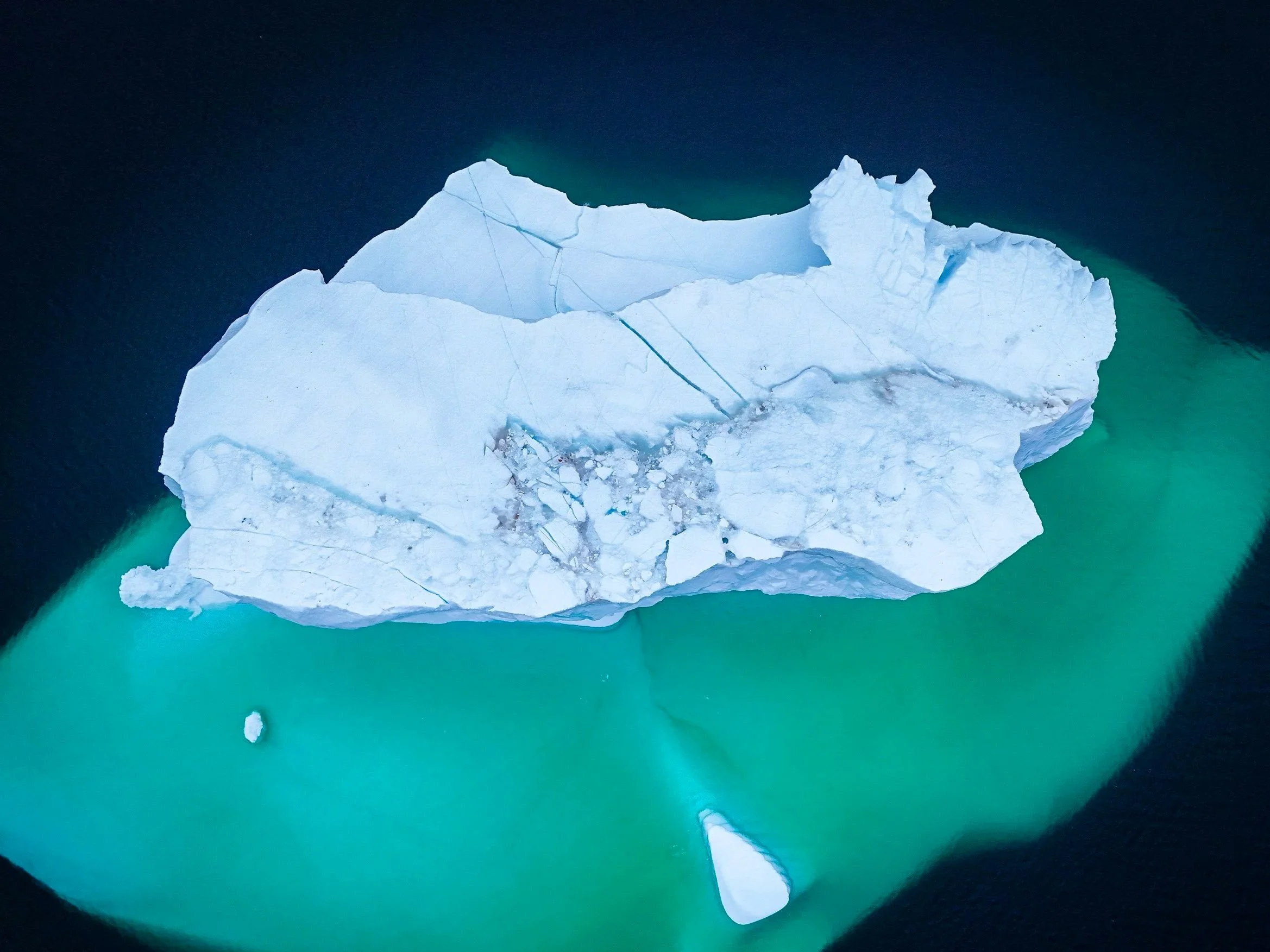A large iceberg floating in dark blue water, with shallower turquoise water around it. The iceberg is irregularly shaped with visible cracks and a small detached piece nearby.