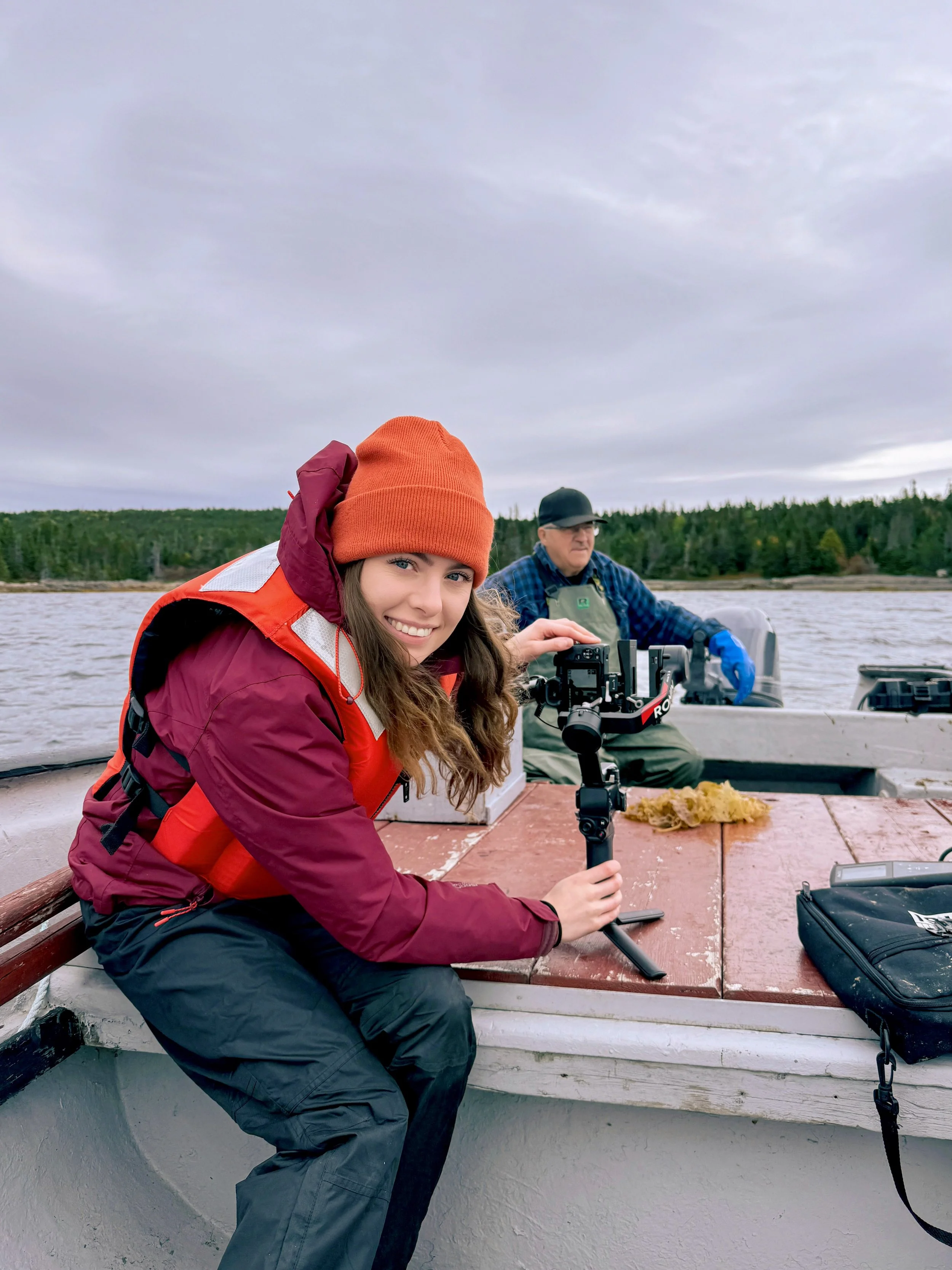 A young woman in outdoor gear on a boat, holding a camera stabilizer, smiling at the camera, with a man in the background operating a boat's motor, water and forested shoreline visible.
