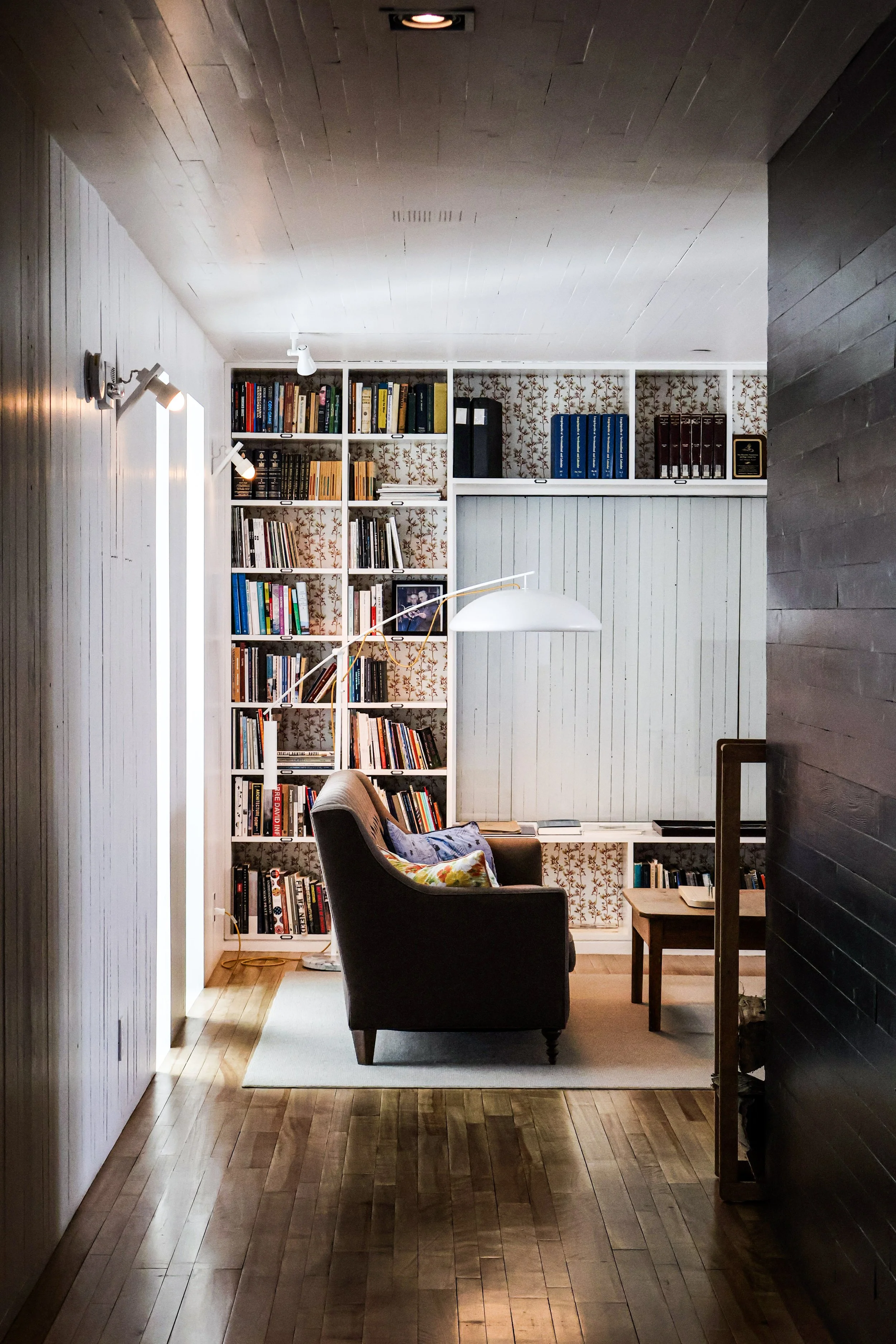 Living room with a white bookshelf filled with books, a brown armchair with cushions, a side table, a white hanging lamp, and a bookshelf behind the armchair, all on a wooden floor.