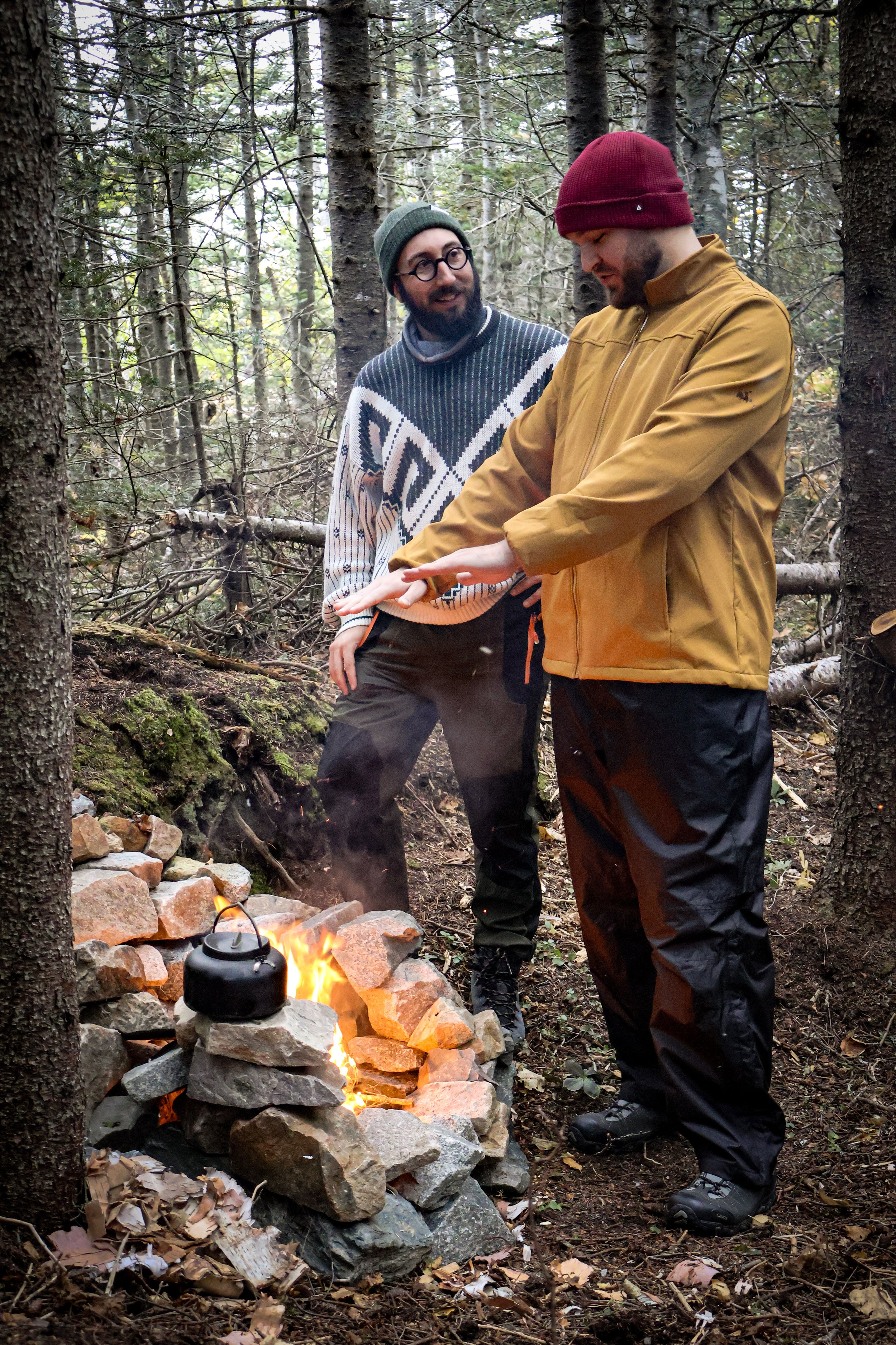 Two men standing in a forest, one is wearing a yellow jacket and red beanie, the other wearing a patterned sweater and glasses, as they tend to a campfire with a black kettle on it.