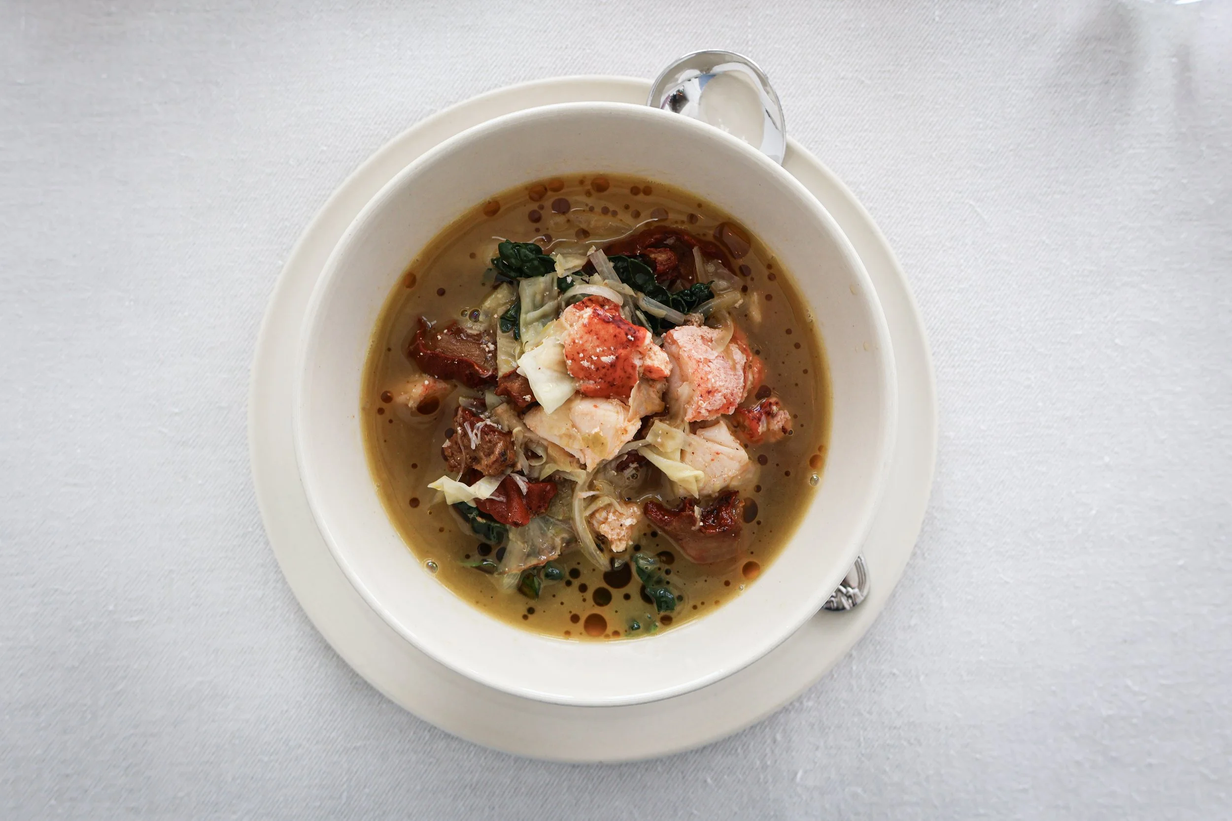 A bowl of seafood soup with chunks of lobster, vegetables, and herbs, served on a white plate with a spoon on the side.
