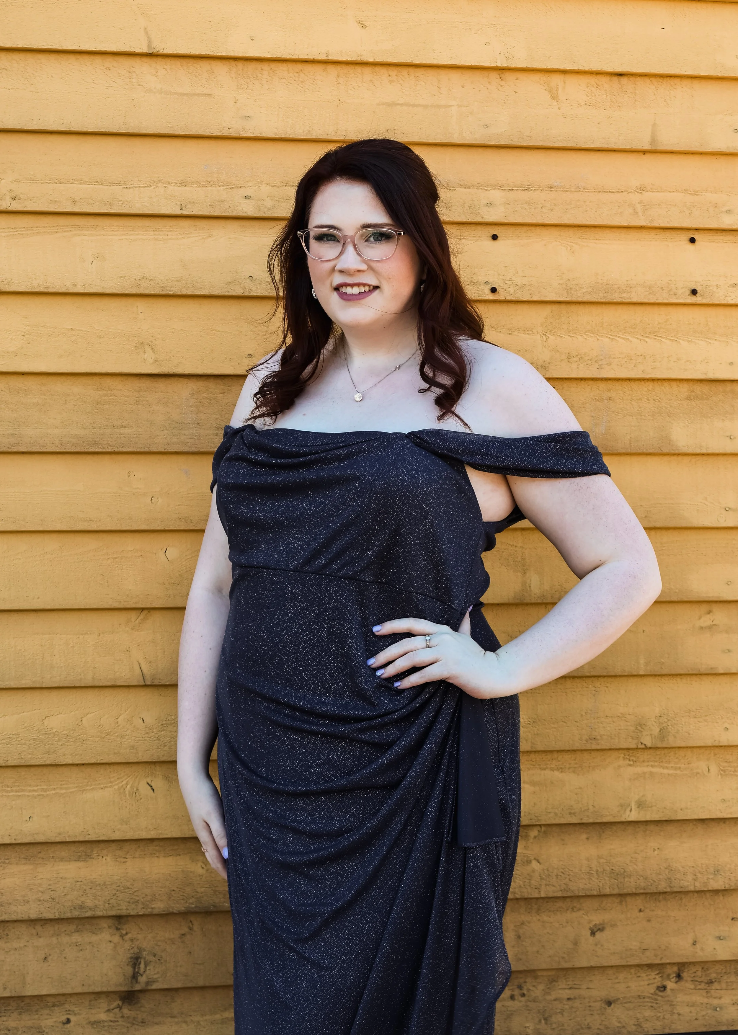 A woman with brown hair, glasses, and earrings standing against a yellow wooden wall, wearing a black off-the-shoulder dress with a side slit, smiling at the camera.