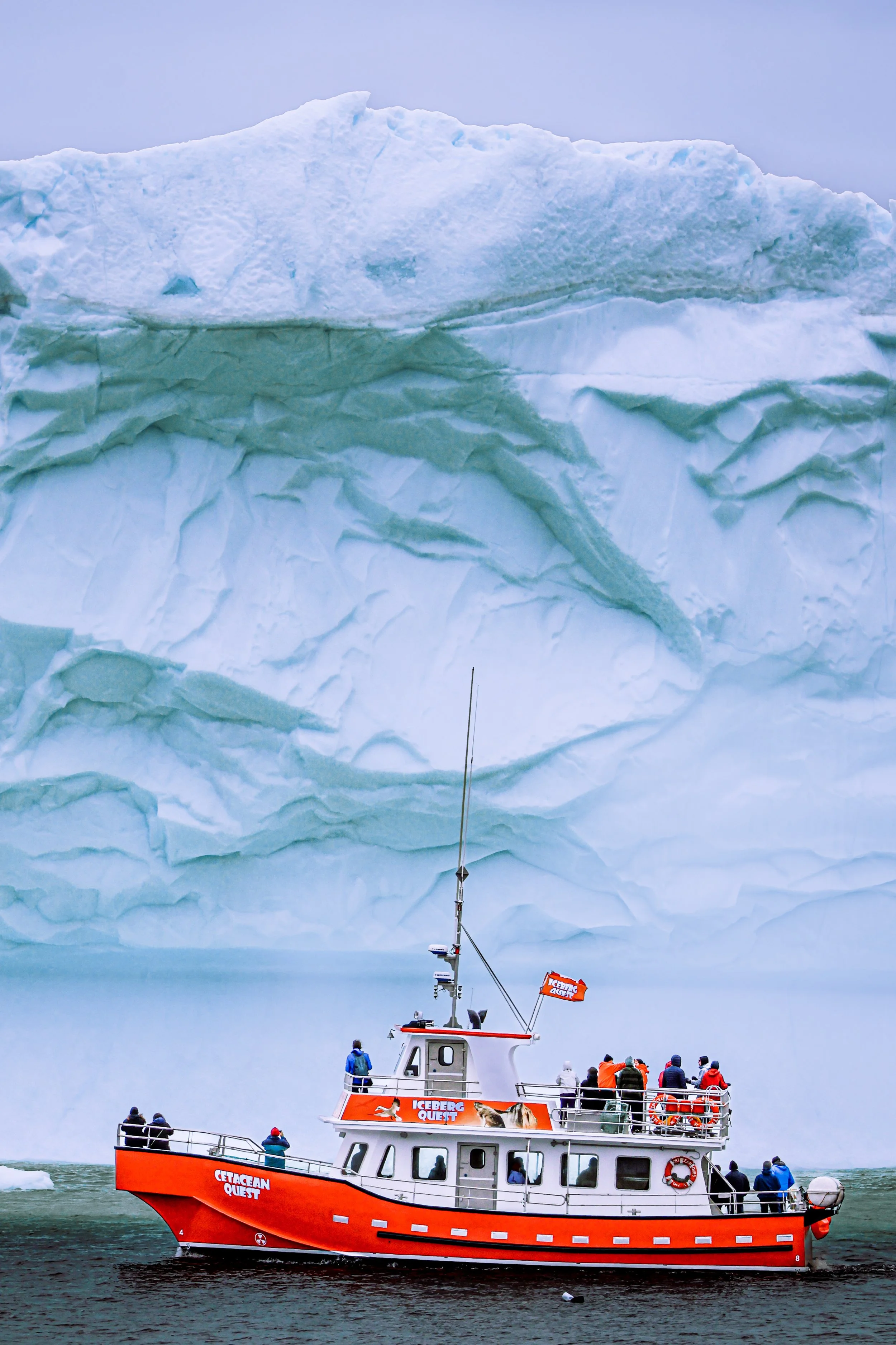 A red boat labeled 'CETACEAN QUEST' with people on board, sailing near a large iceberg with deep crevices.