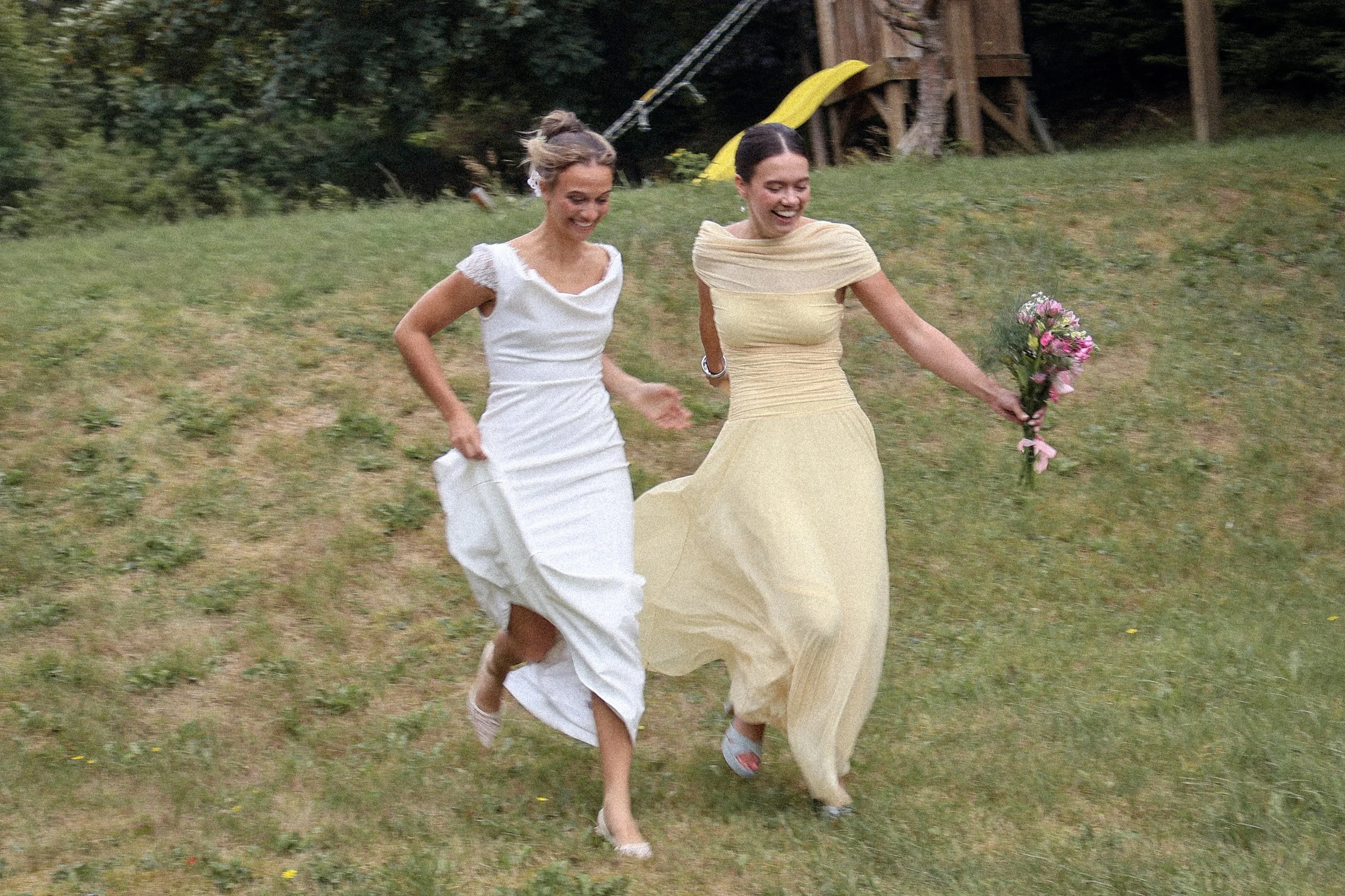 Two women in dresses running on a grassy hill, one holding a bouquet of pink flowers, with a slide and wooden structure in the background.