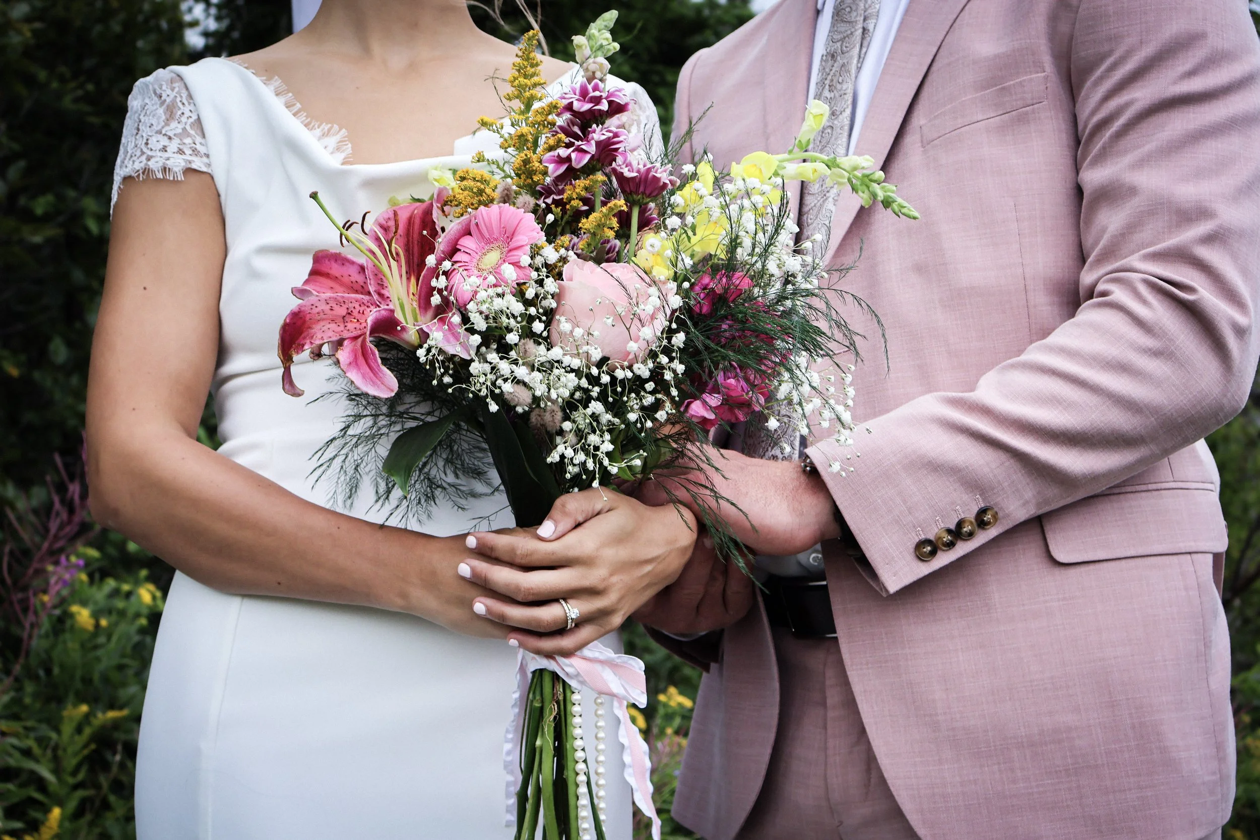 A bride holding a bouquet of pink, white, and yellow flowers, standing next to a groom in a pink suit, outdoors surrounded by greenery.